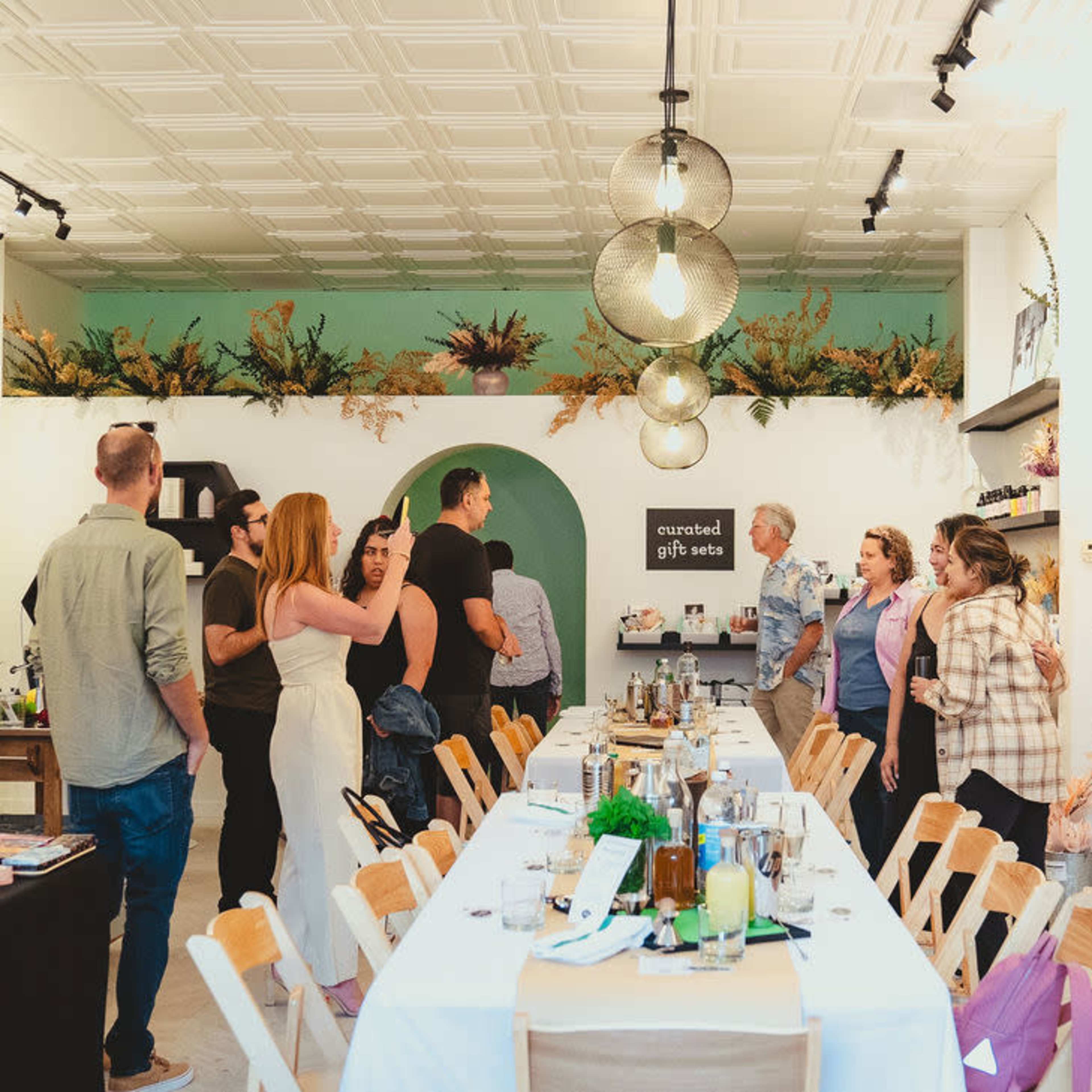 A group of people stands around a long table in a well-decorated shop filled with plants and gift items.