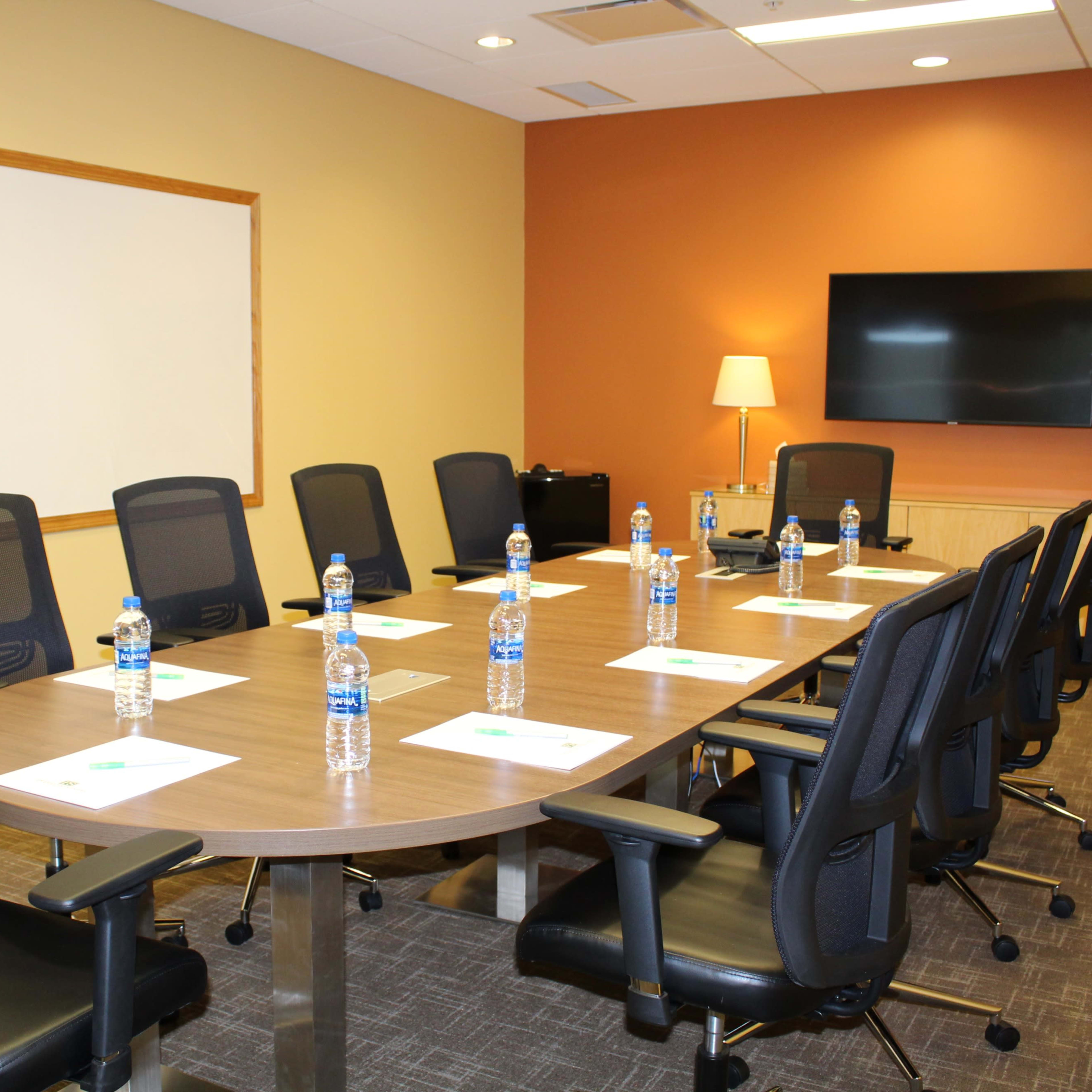 A conference room is set up with a large oval table surrounded by black chairs, each equipped with a water bottle and a notepad, along with a television and a whiteboard on the walls.