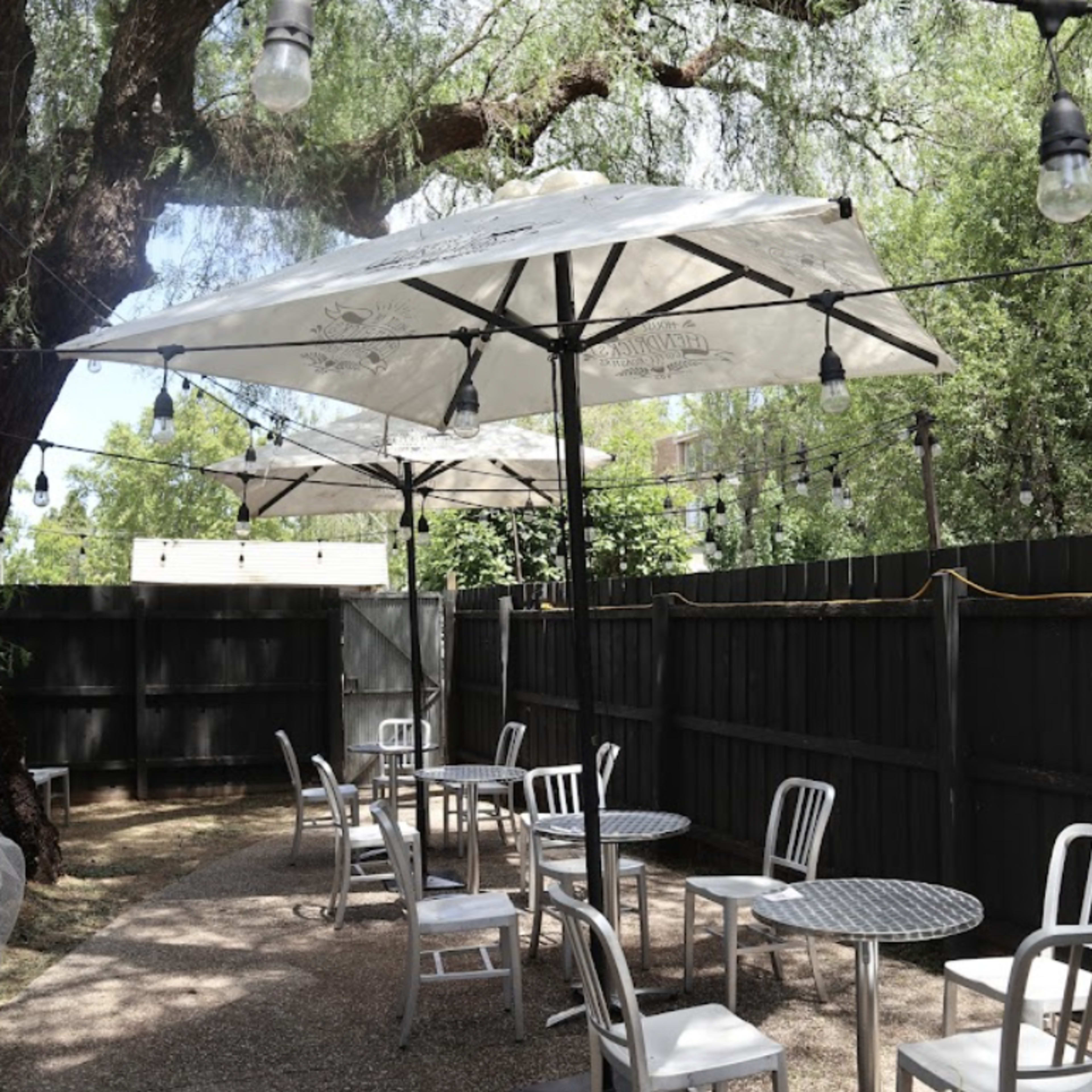 A patio area features several tables with white umbrellas surrounded by a wooden fence and string lights.