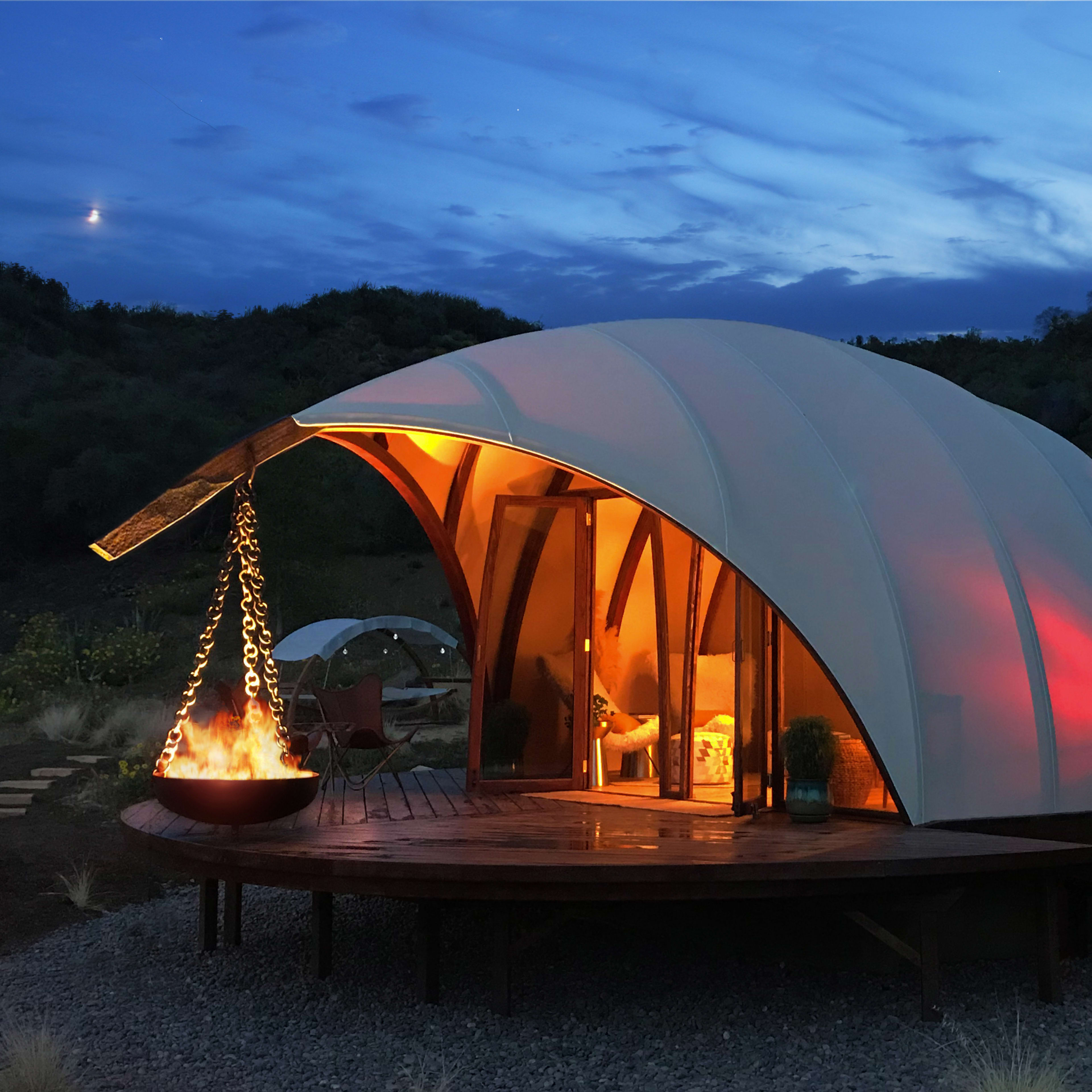 A modern dome-shaped structure with warm lighting is set on a wooden deck surrounded by a rocky landscape at dusk.