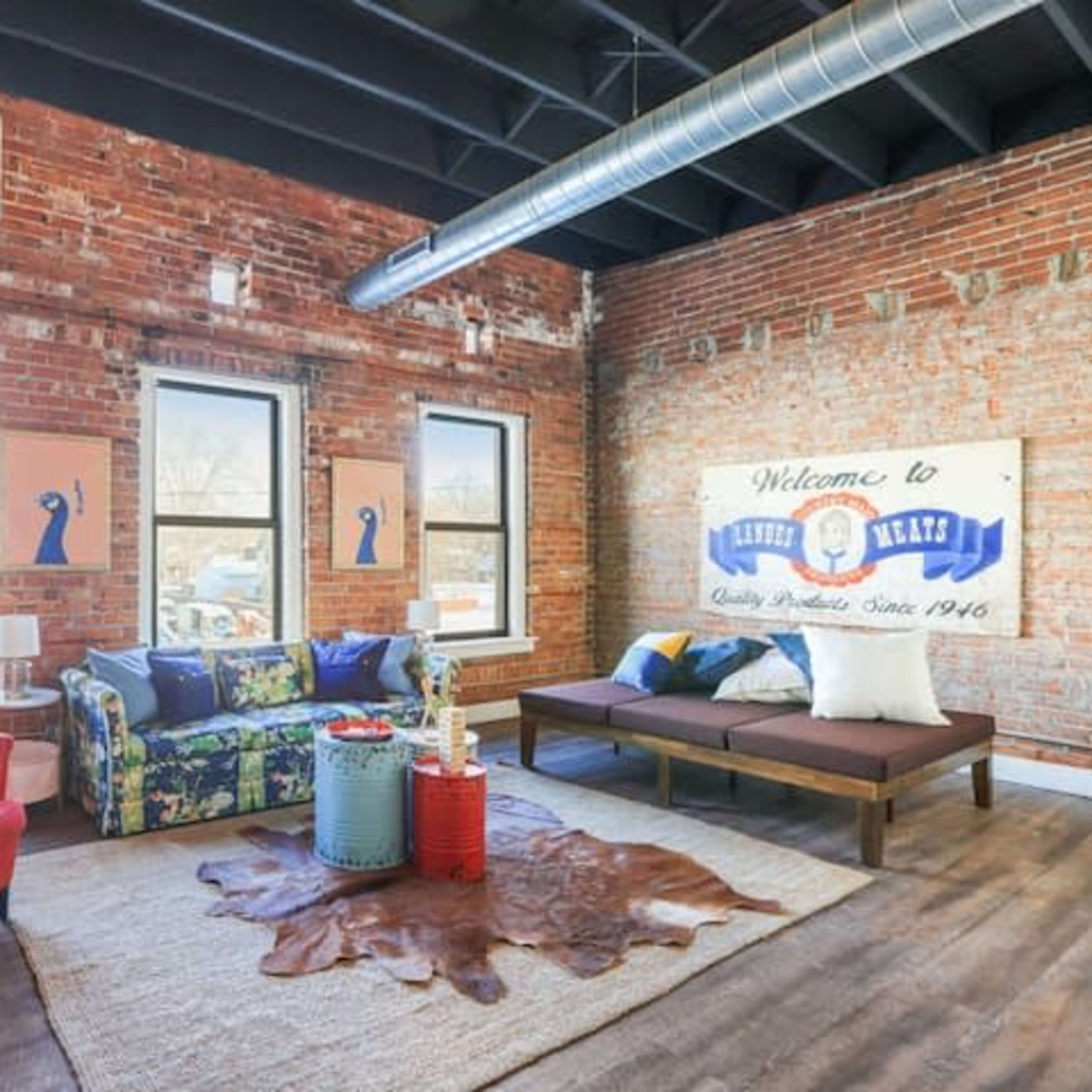The living room features a mix of red and blue furniture, a large area rug, exposed brick walls, and large windows letting in natural light.