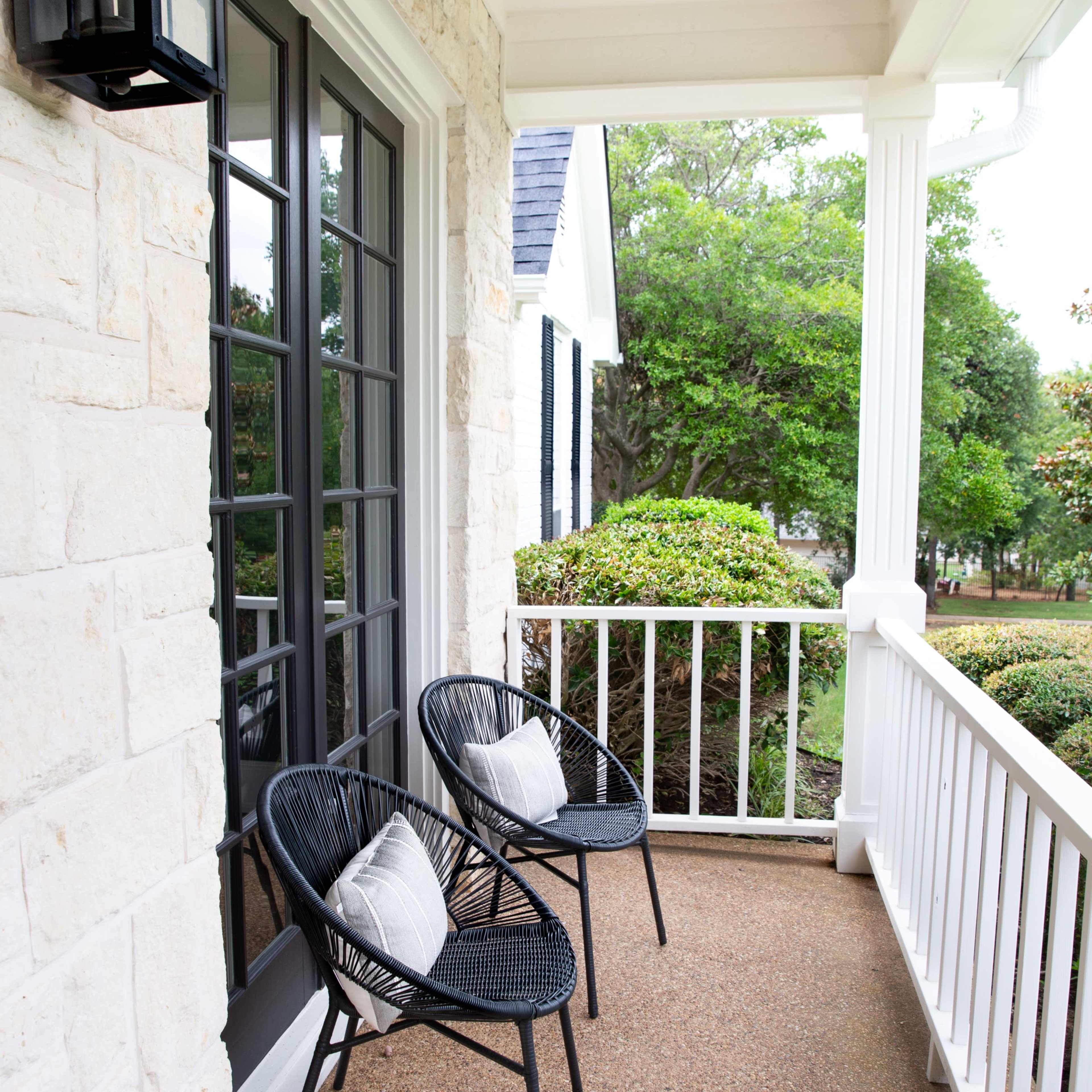 A porch with two black chairs and a small table, surrounded by greenery and a stone wall.