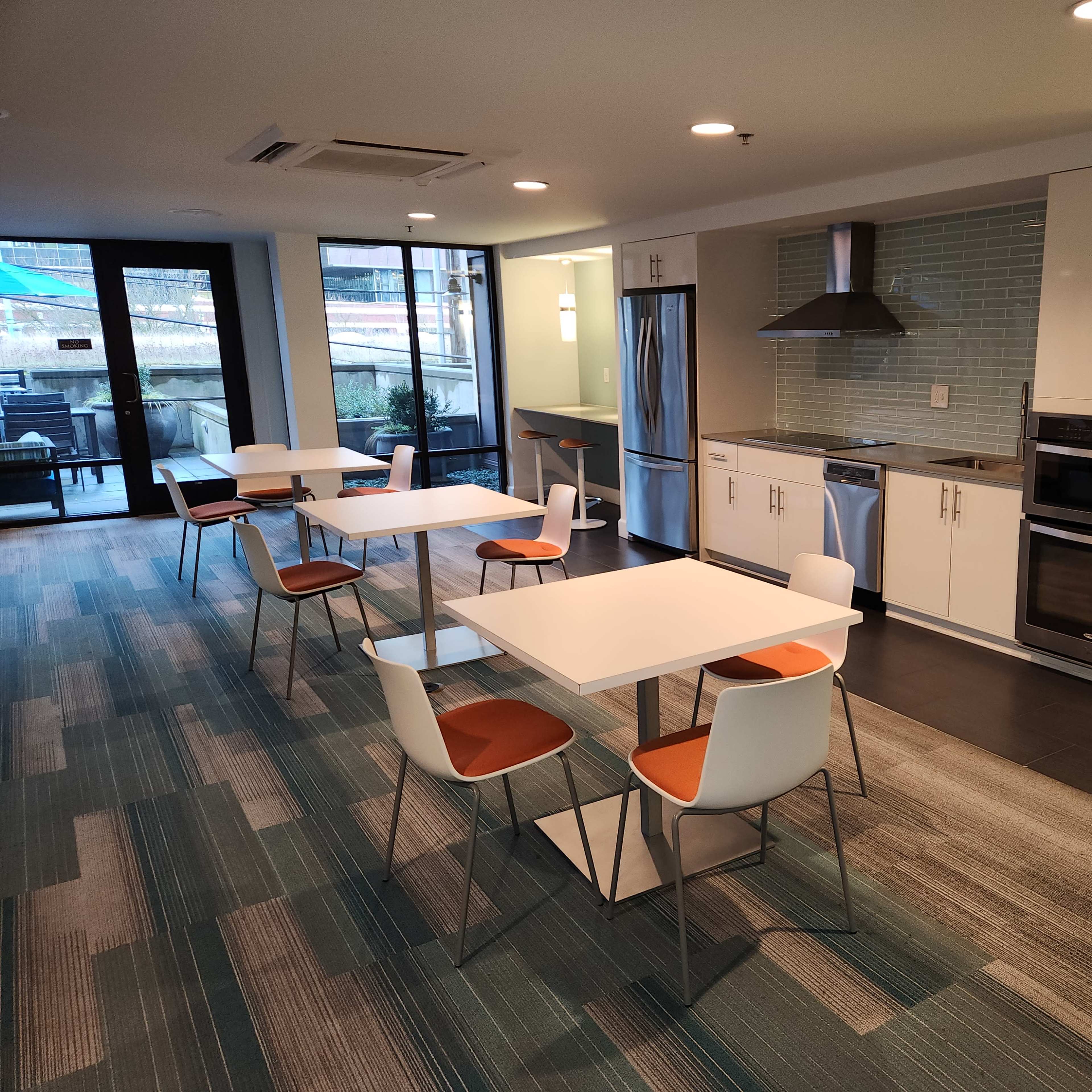 A modern kitchen and dining area featuring stainless steel appliances, white tables with orange-accented chairs, and large windows overlooking an outdoor space.