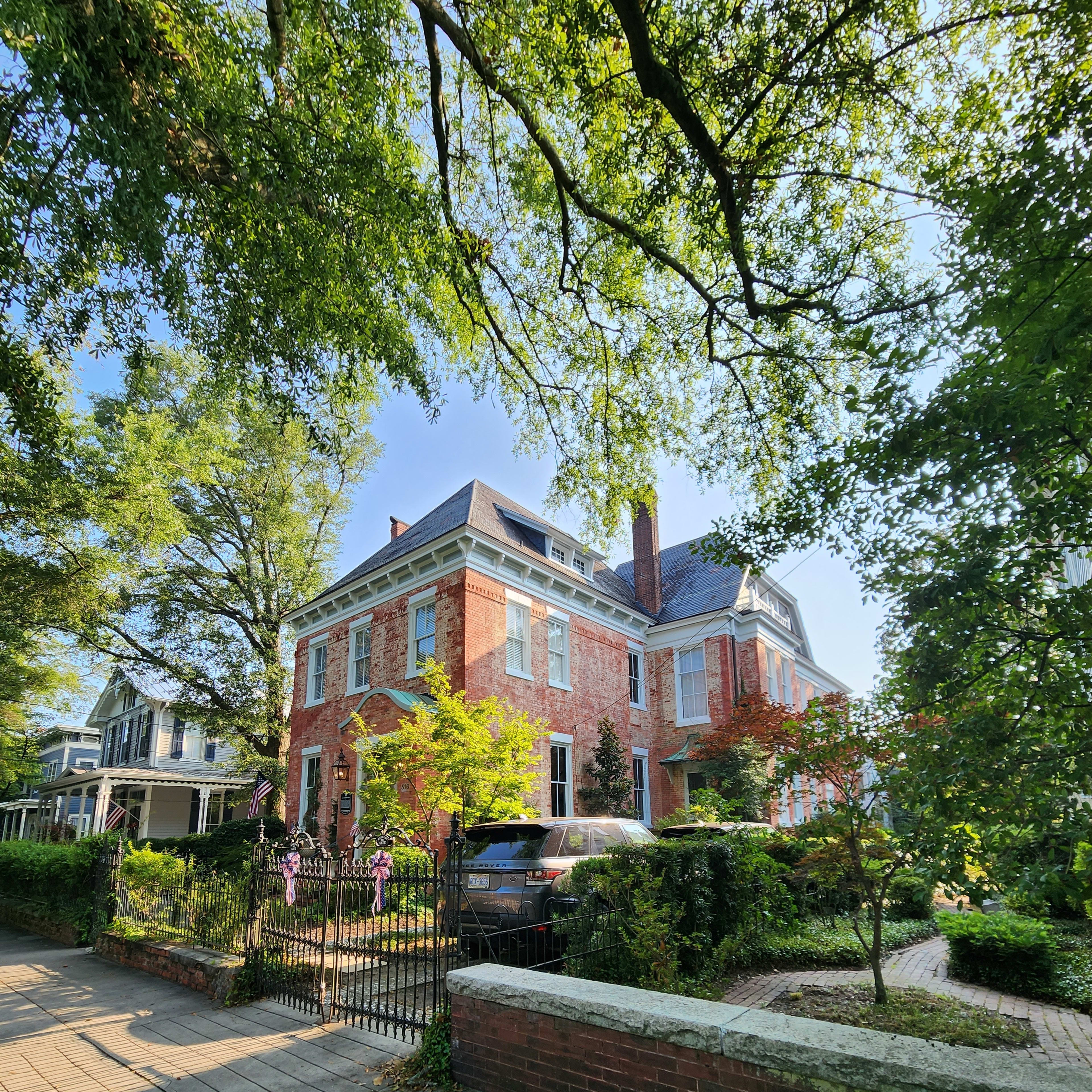 A large red brick house with a steep roof is surrounded by greenery and a wrought iron fence.
