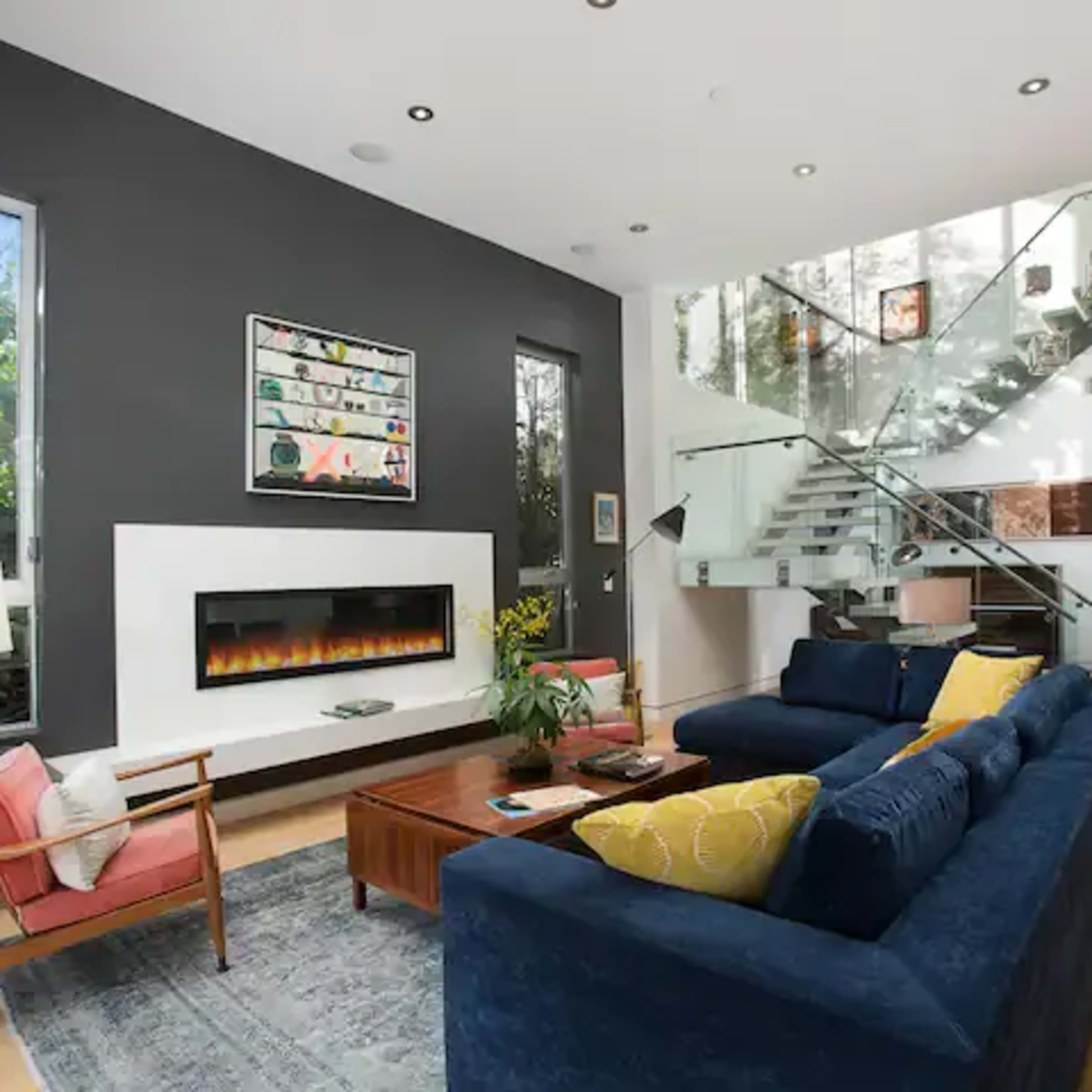 A modern living room with a large blue sofa, a wooden coffee table, and a wall-mounted fireplace, featuring tall windows and a staircase with glass railings.