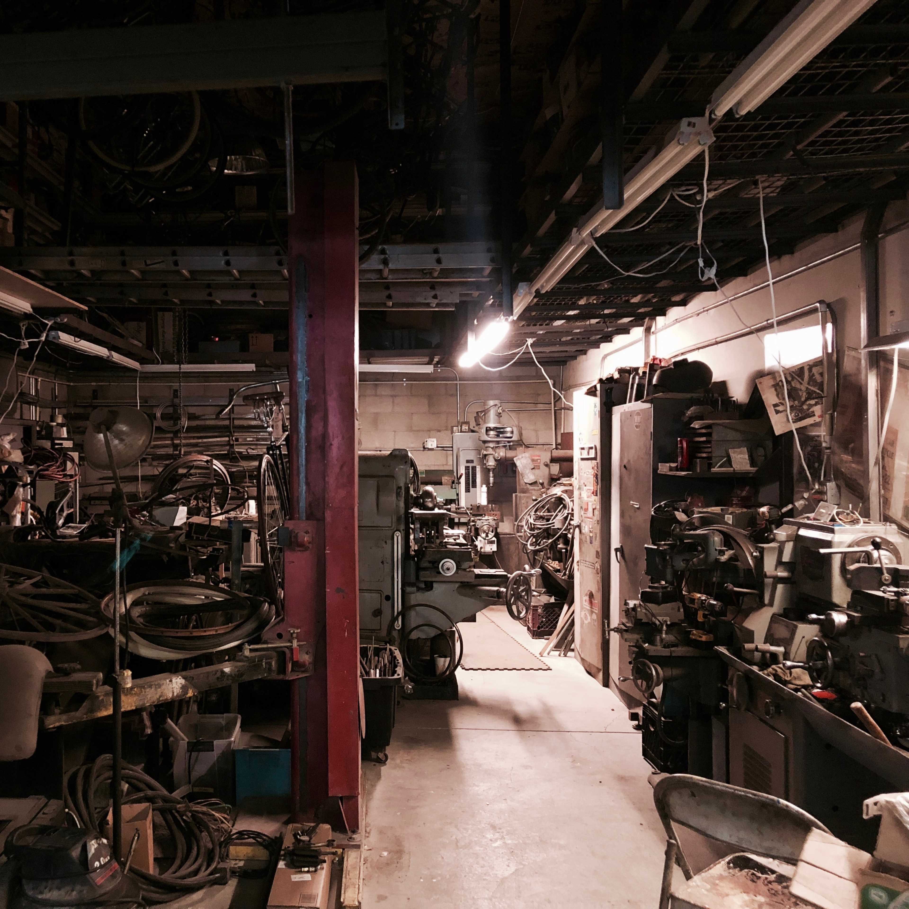 A cluttered workshop filled with various tools, machinery, and remnants of bicycles in dim lighting.