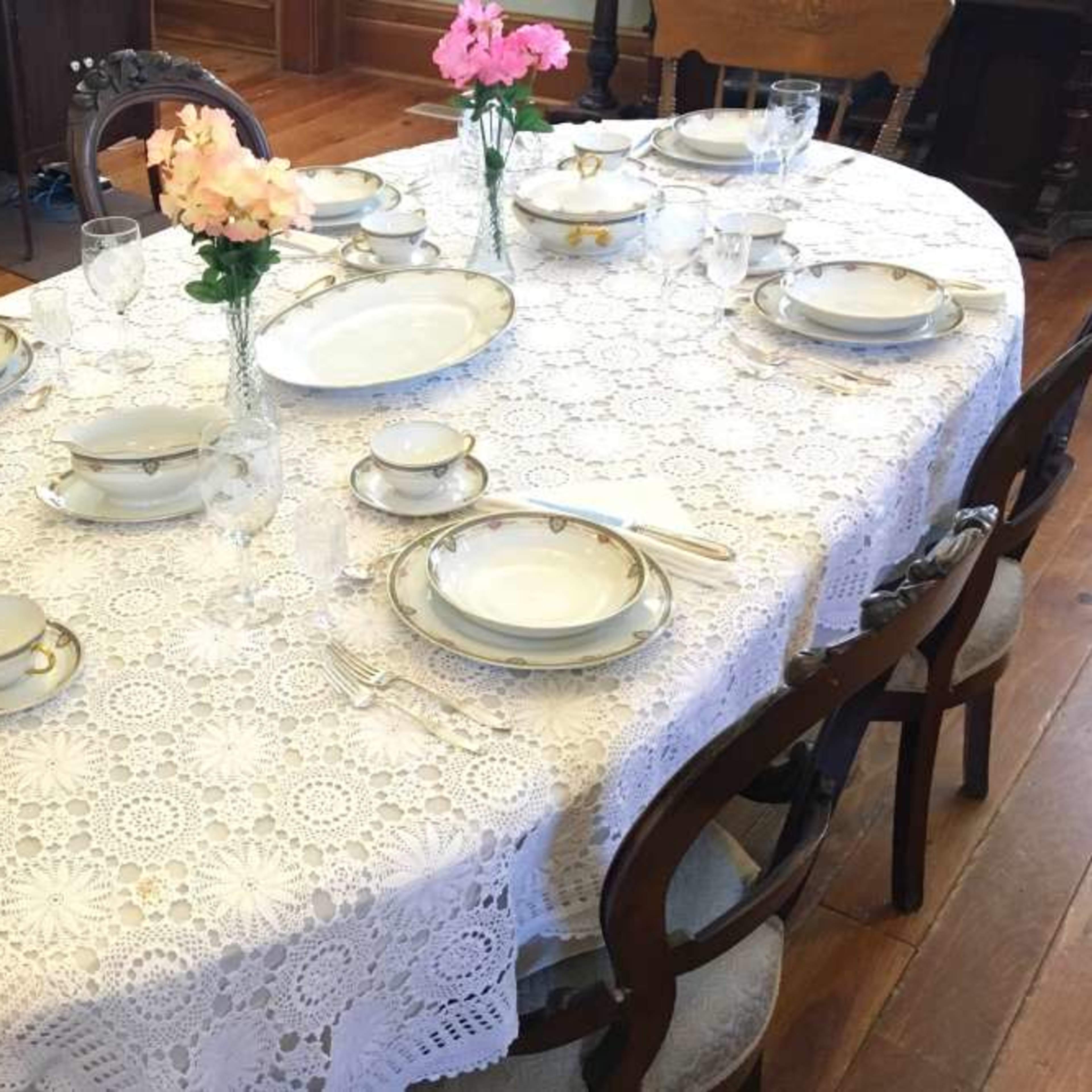 A dining table is set with elegant plates, cutlery, and floral centerpieces, covered by a lace tablecloth.