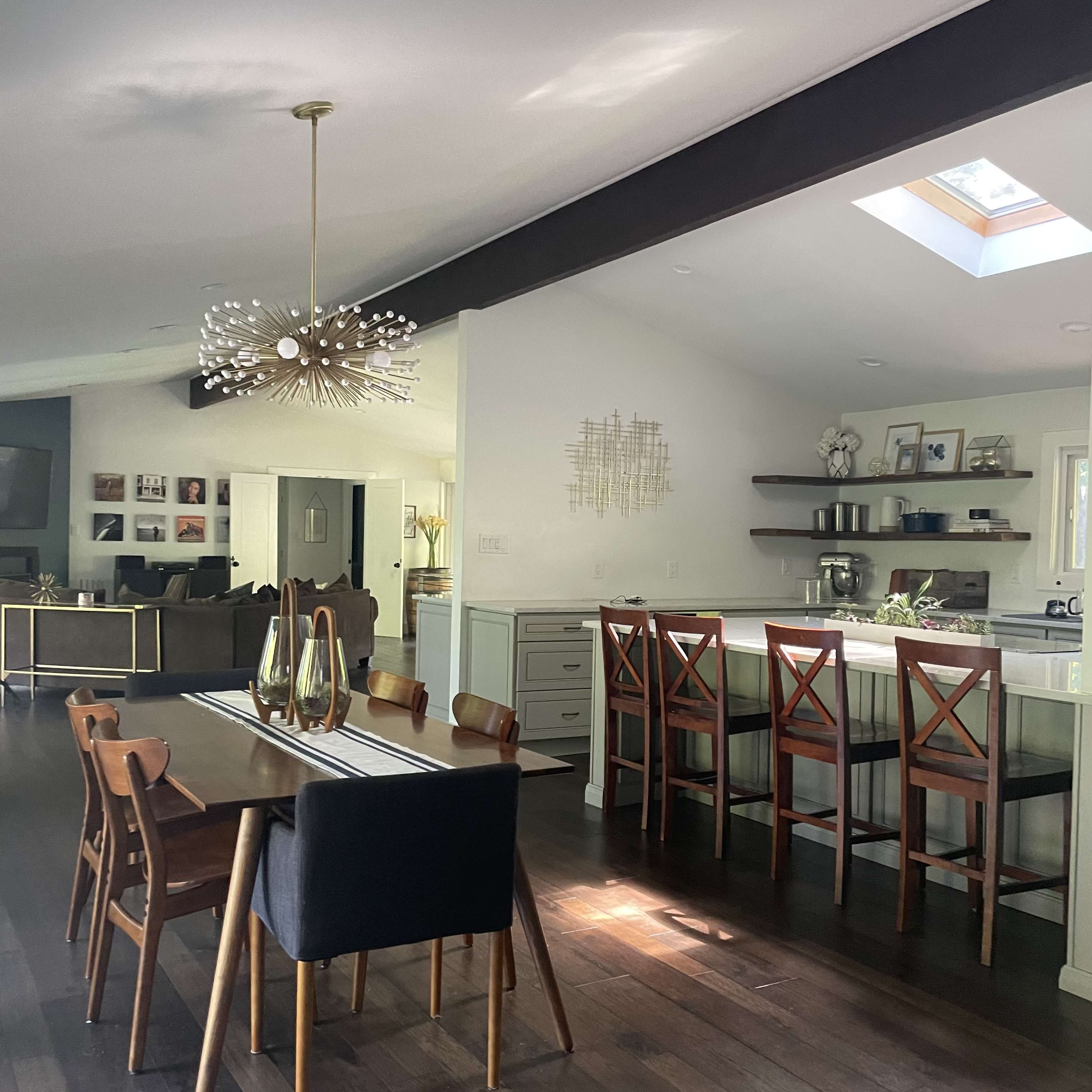 A dining area with a wooden table and chairs is adjacent to a kitchen with bar seating, all illuminated by natural light through a skylight.