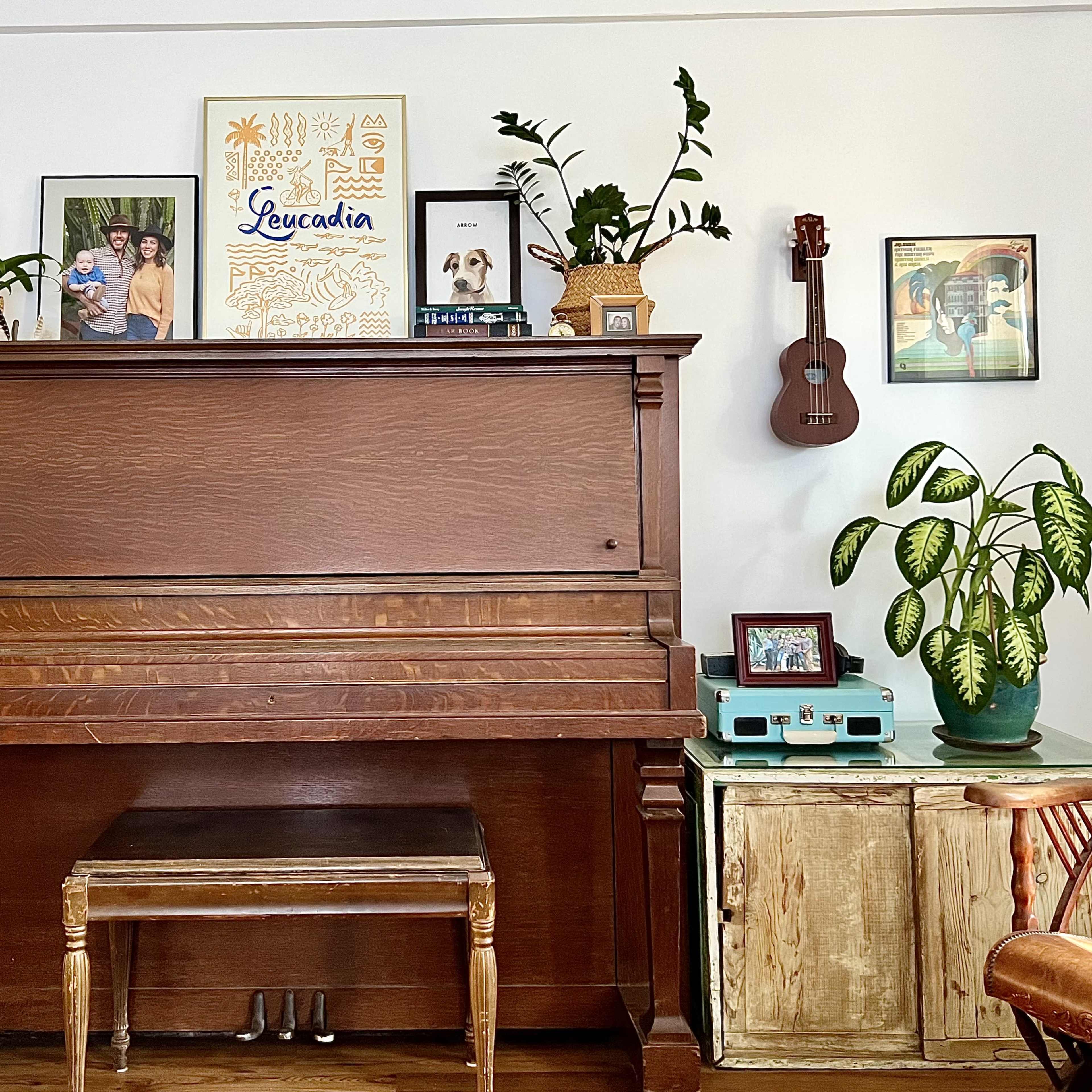 A wooden piano with a decorative vase of plants and framed photos on the wall behind it, alongside a ukulele and a green storage cabinet.