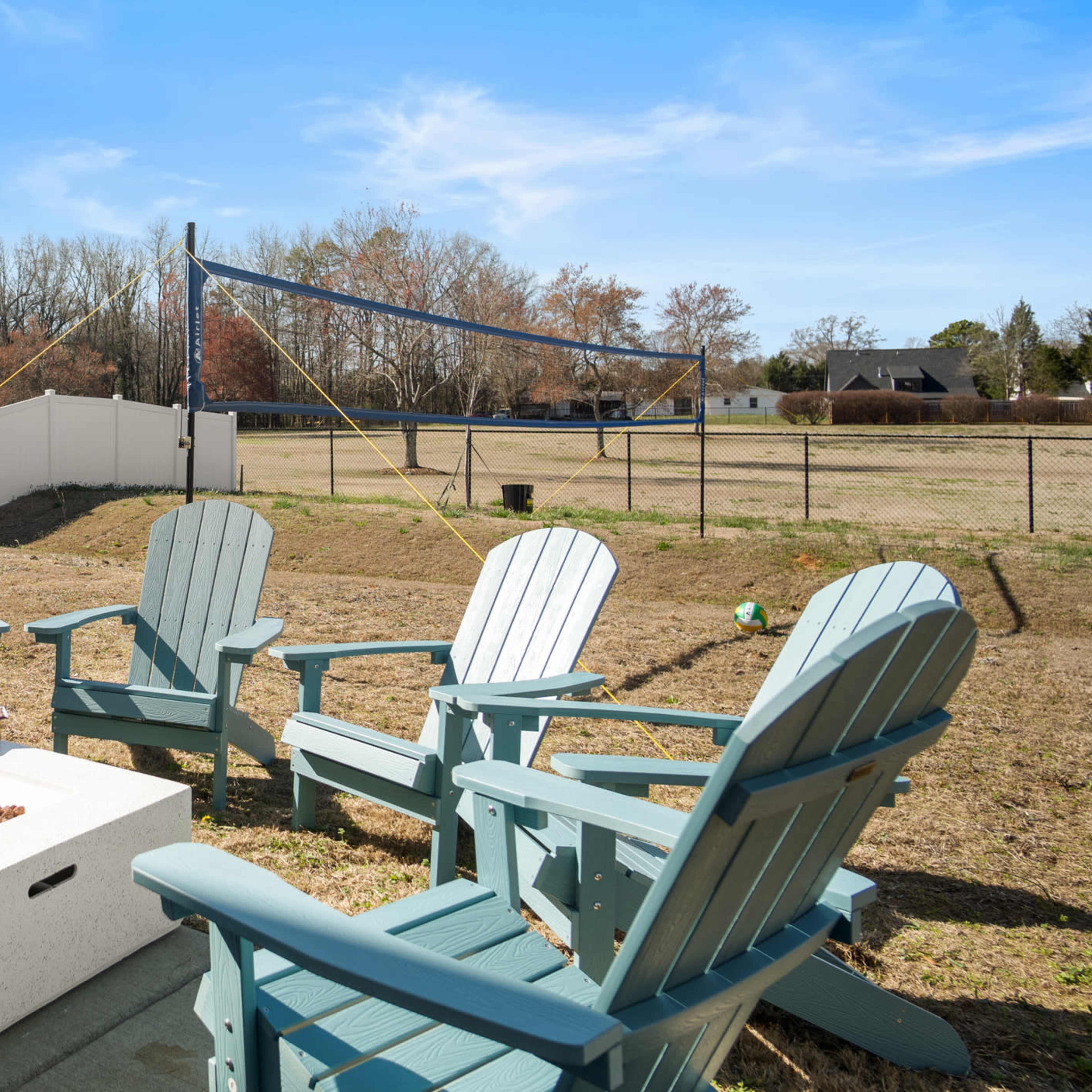 A backyard scene with teal Adirondack chairs arranged around a fire pit, set against a fenced area with a volleyball net in the background.