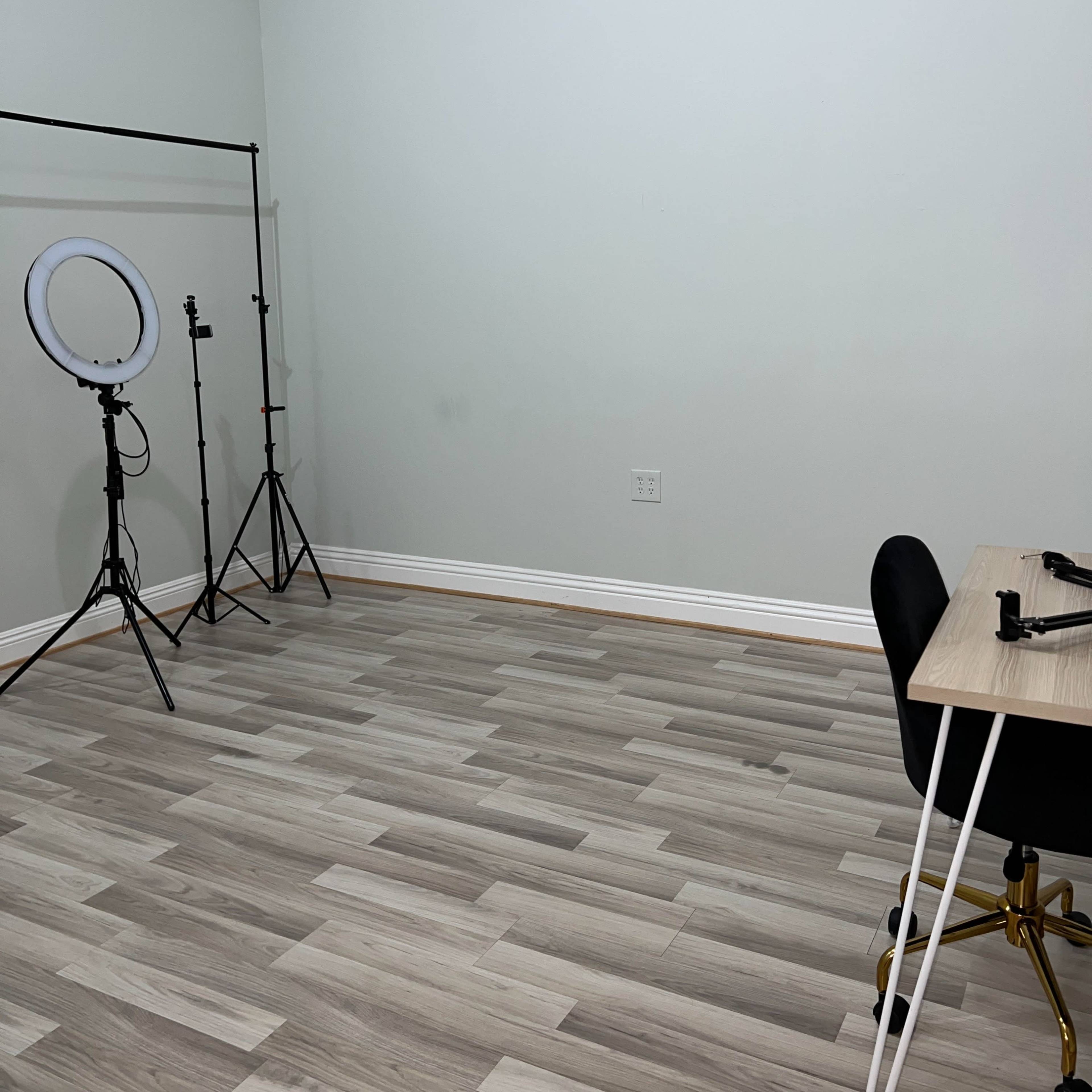 A minimalist room with light wood flooring, a desk with a black chair, and equipment stands for lighting or photography against a plain wall.