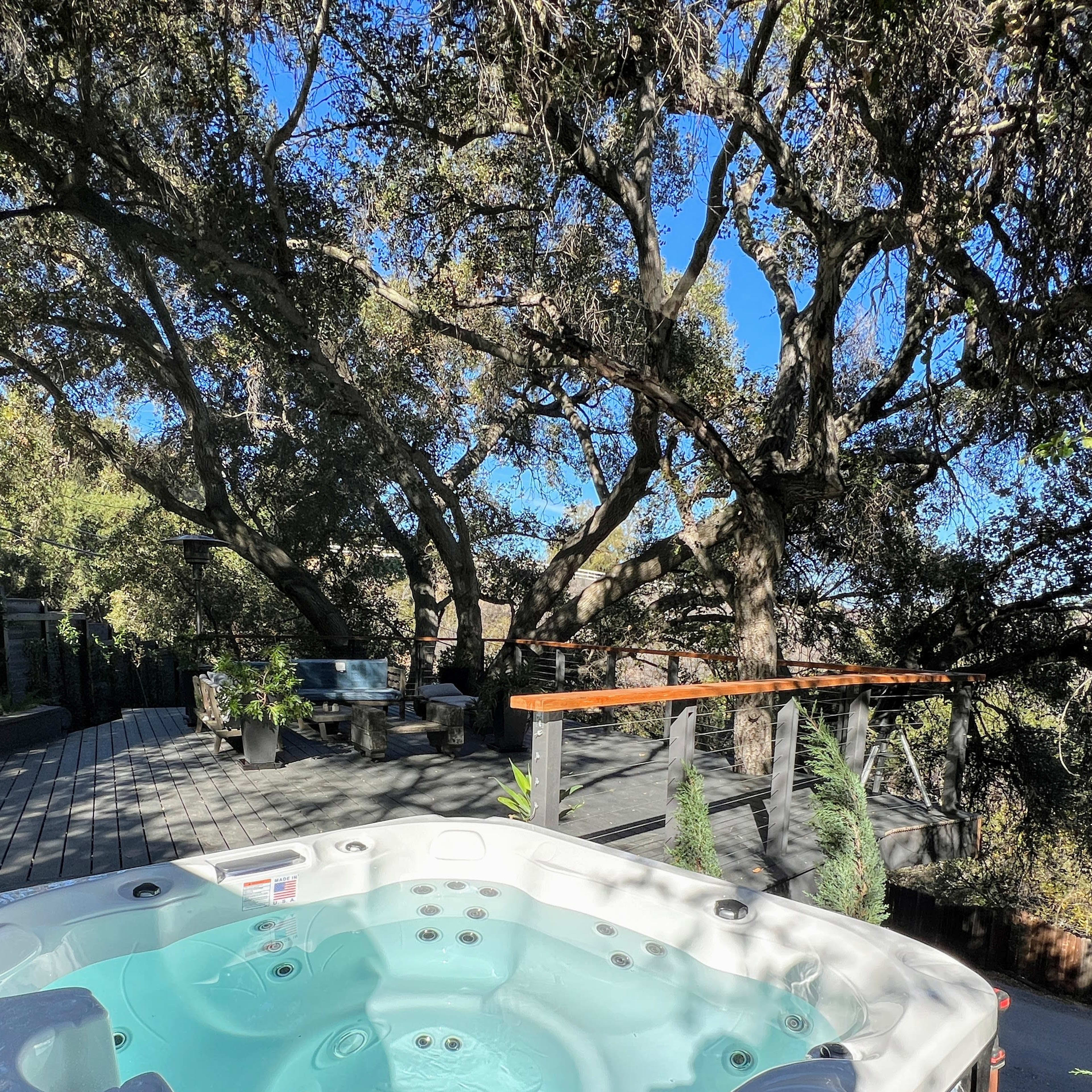 A hot tub is positioned on a deck surrounded by large trees and a shaded seating area.