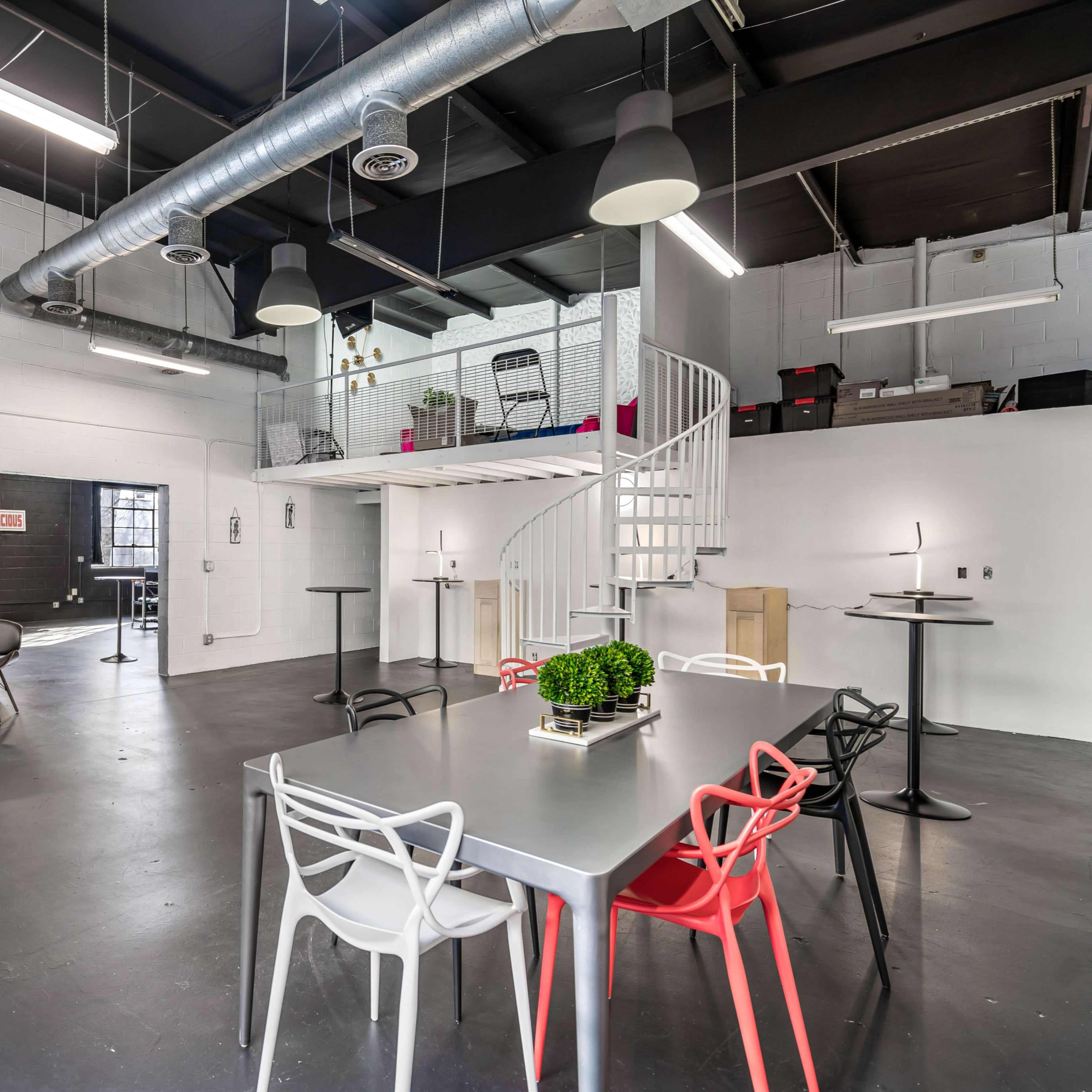 A spacious, modern interior with a mix of seating arrangements, including a blue sofa, a metallic table with colorful chairs, and a spiral staircase leading to a mezzanine level.