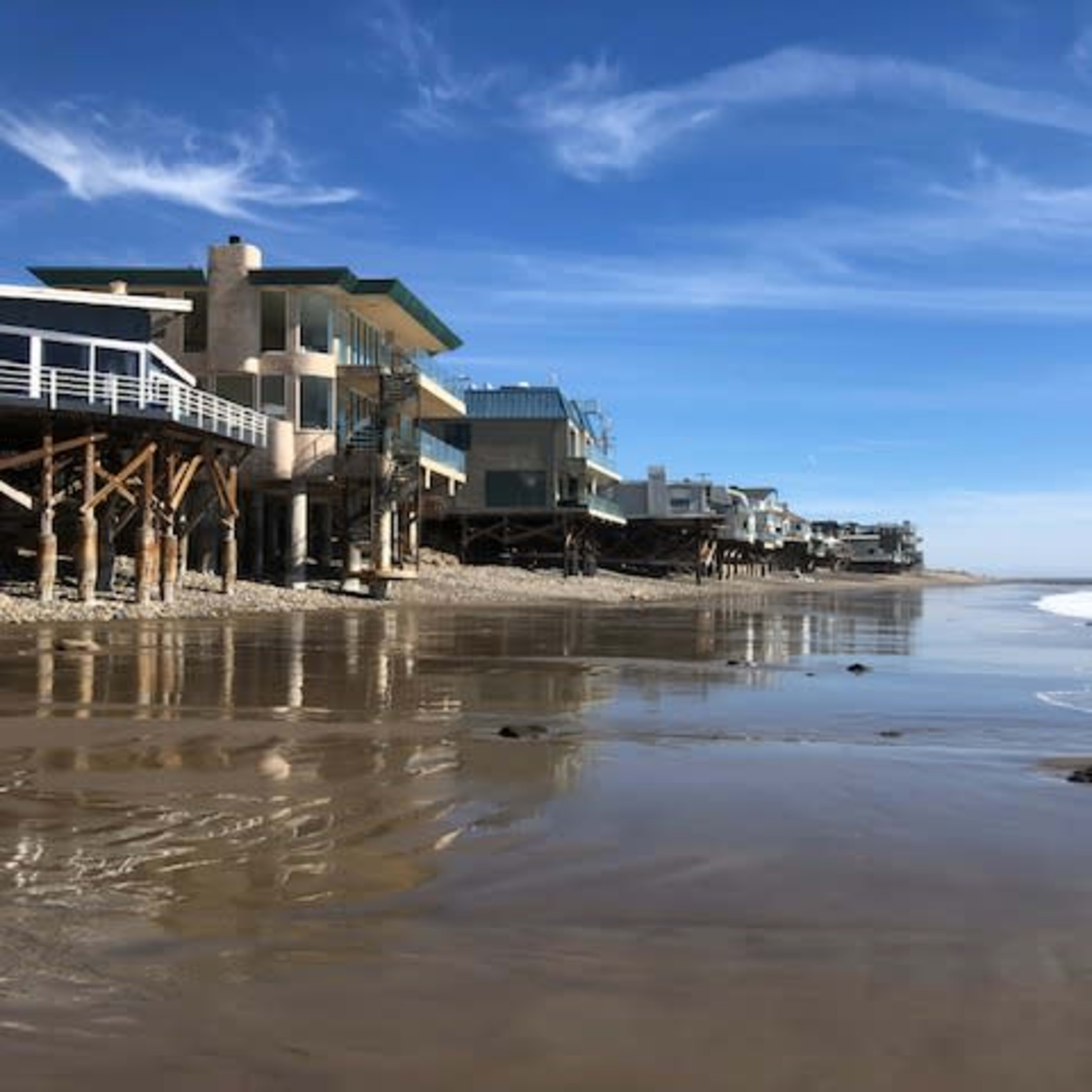 A row of beach houses built on stilts along a sandy shoreline, with gentle waves lapping at the beach.