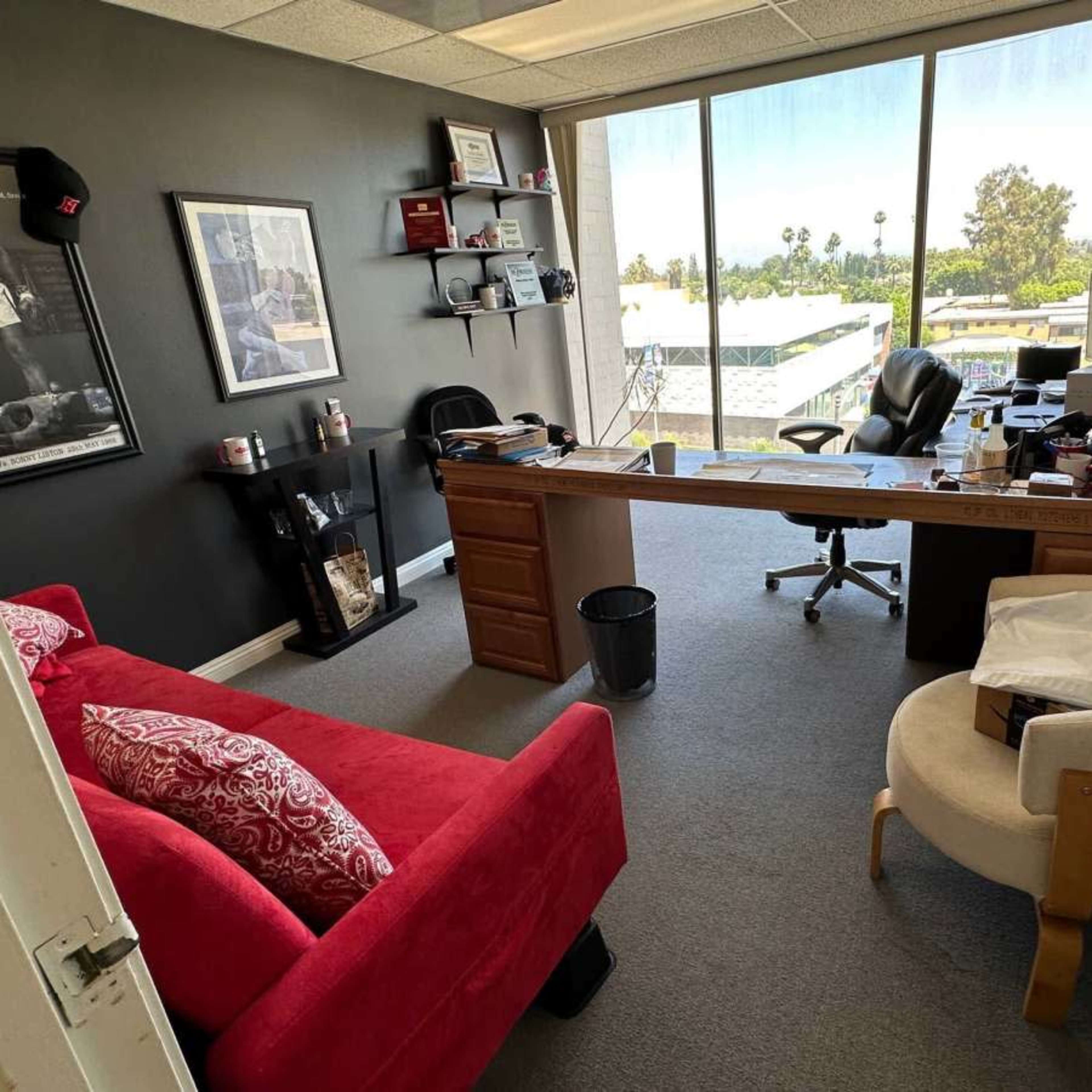 A modern office with a large window, a desk with a chair, and a red sofa next to a white armchair.
