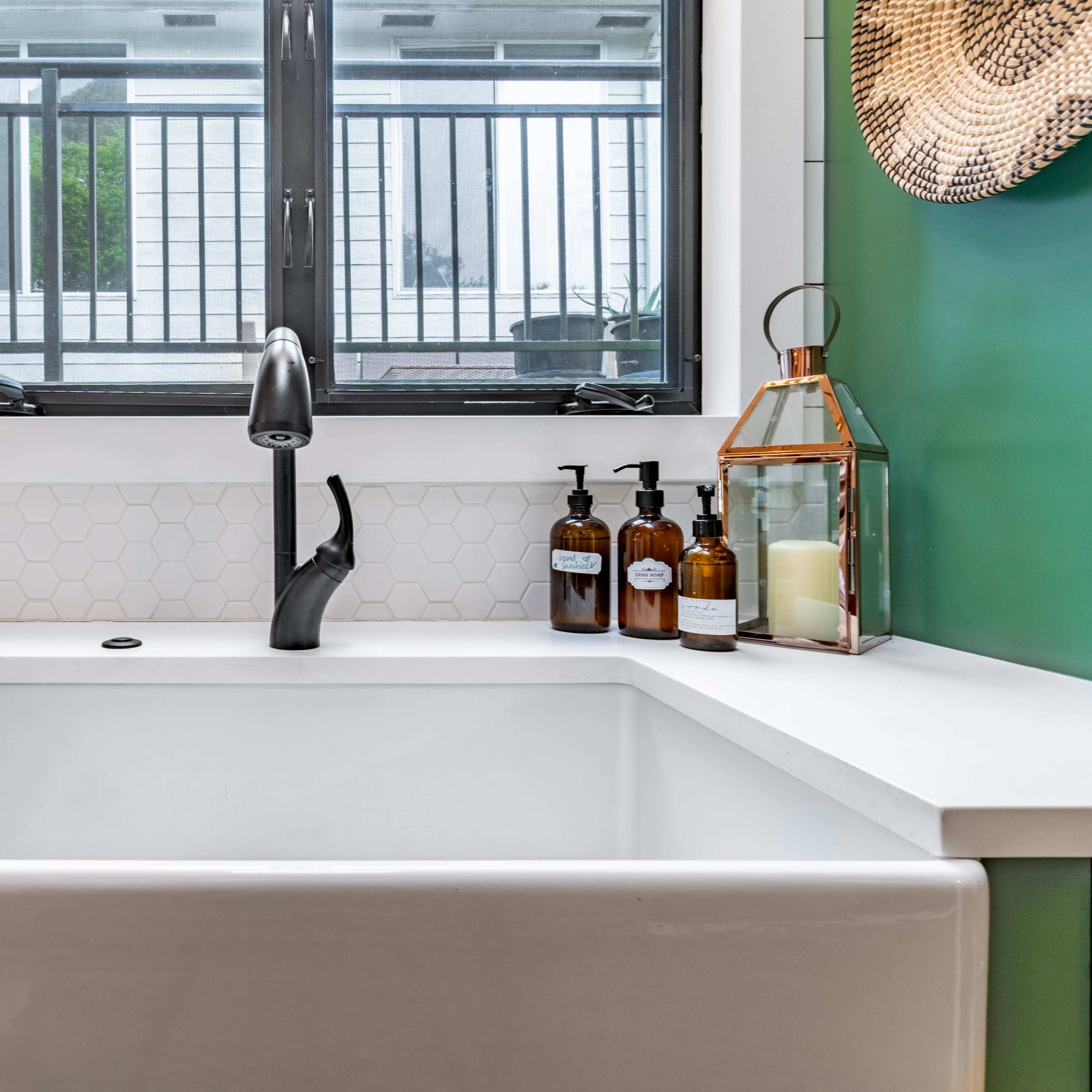 A modern kitchen sink area featuring a farmhouse-style sink, a sleek black faucet, and decorative items including a plant, amber bottles, and a lantern on a green countertop.