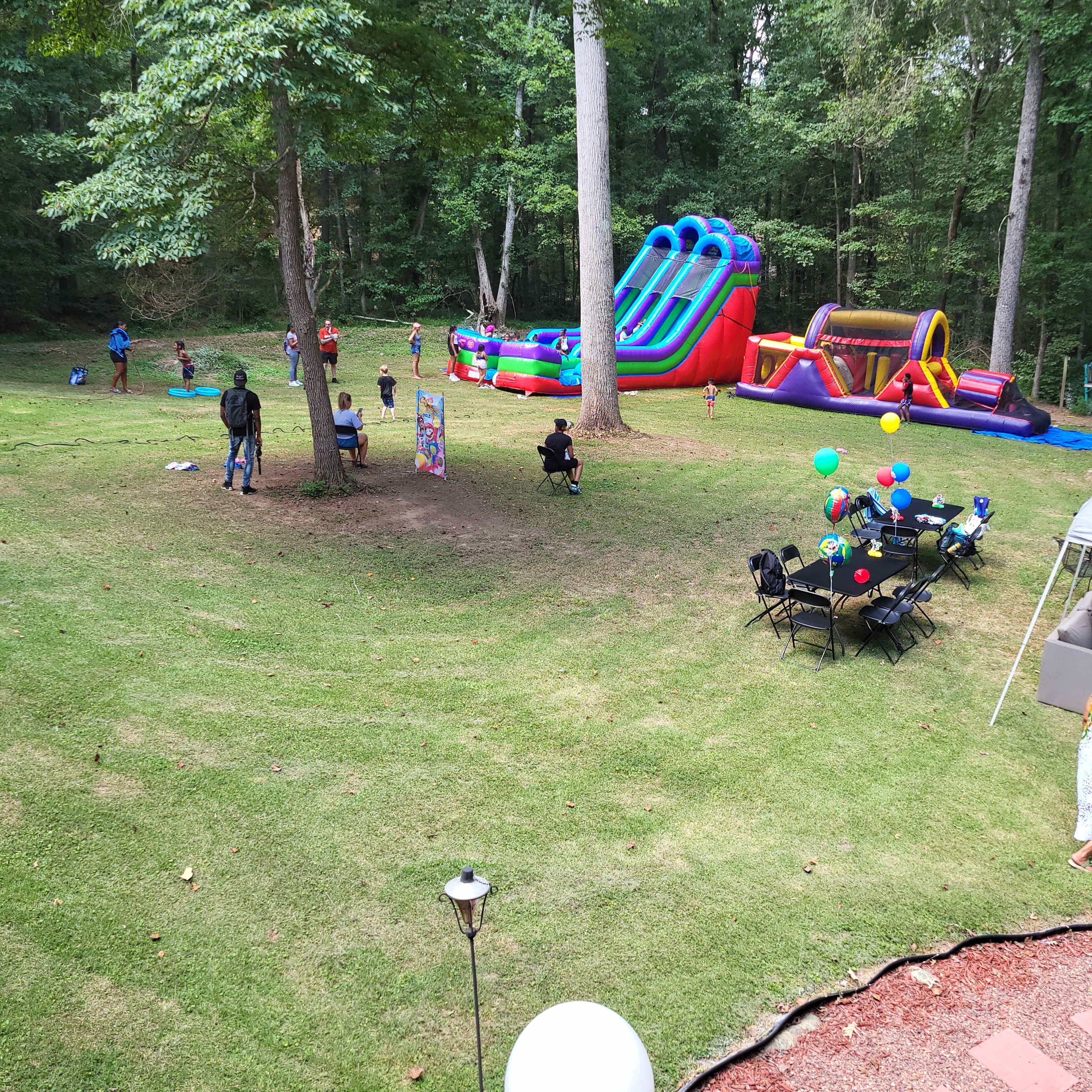 A backyard with inflatable slides and games, children playing, and tables set with balloons and chairs.