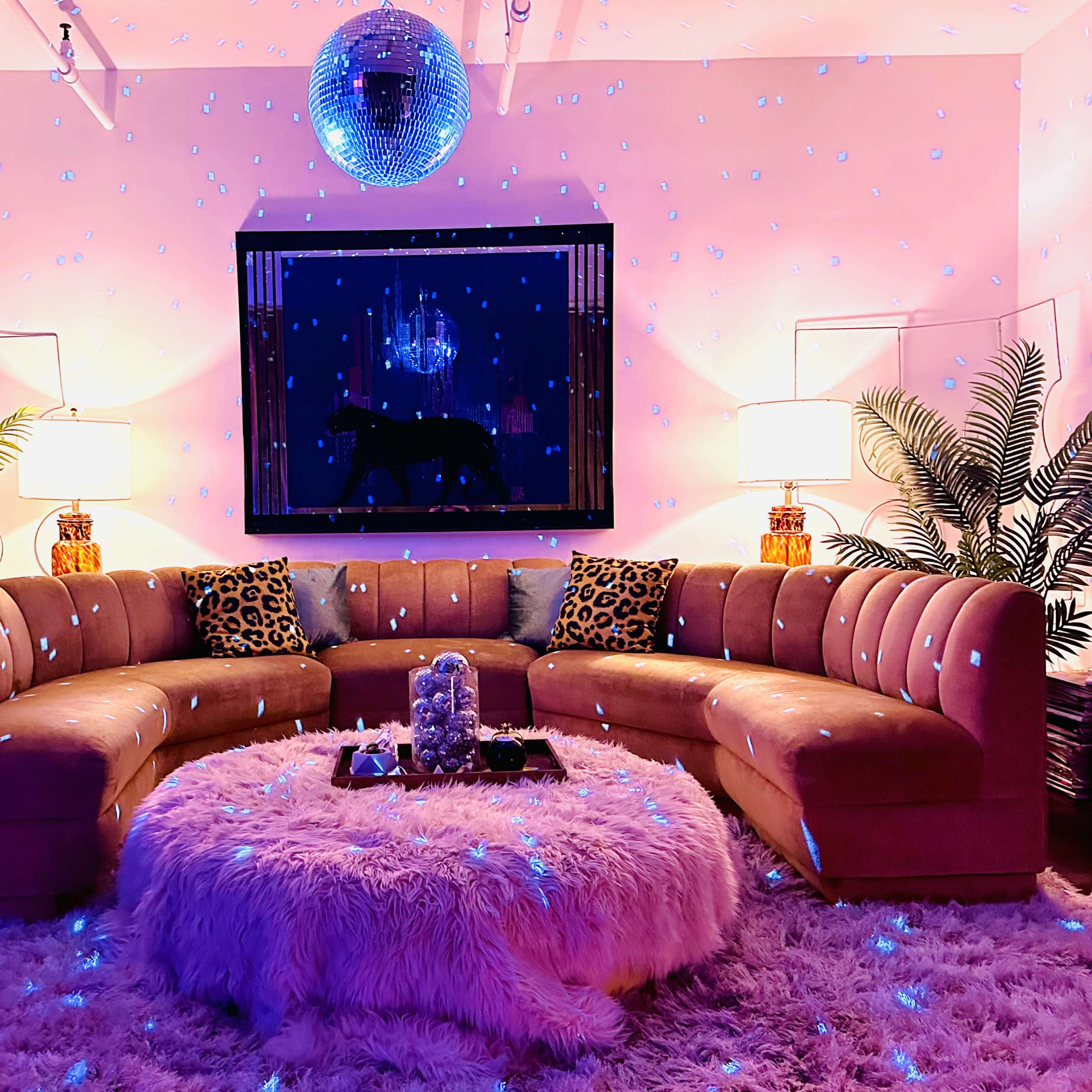 A spacious living room features a curved velvet sofa, a round shag rug, and disco ball lighting, with palm plants and framed artwork on the walls.