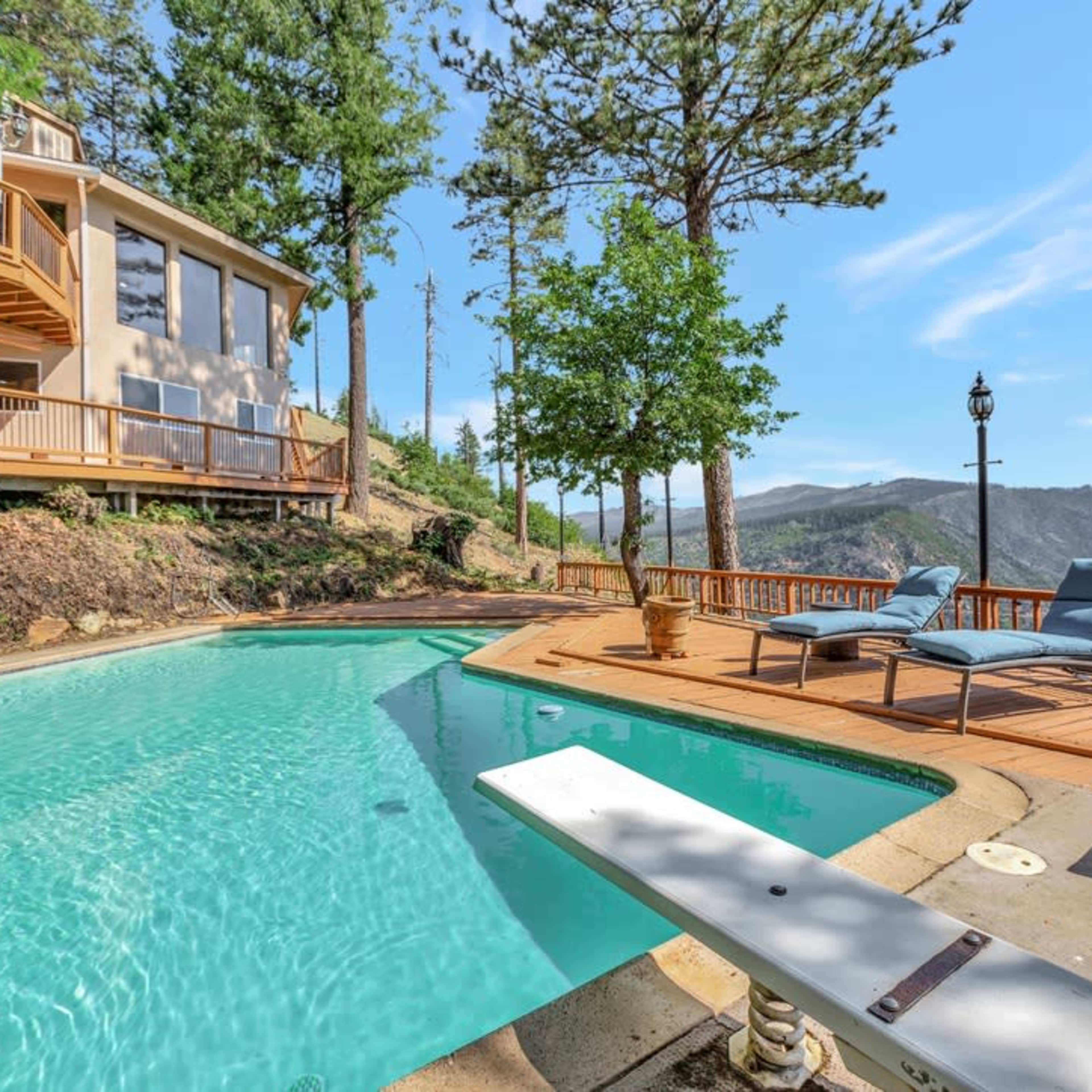 A clear swimming pool sits beside a wooden deck, surrounded by trees and overlooking a mountainous landscape.