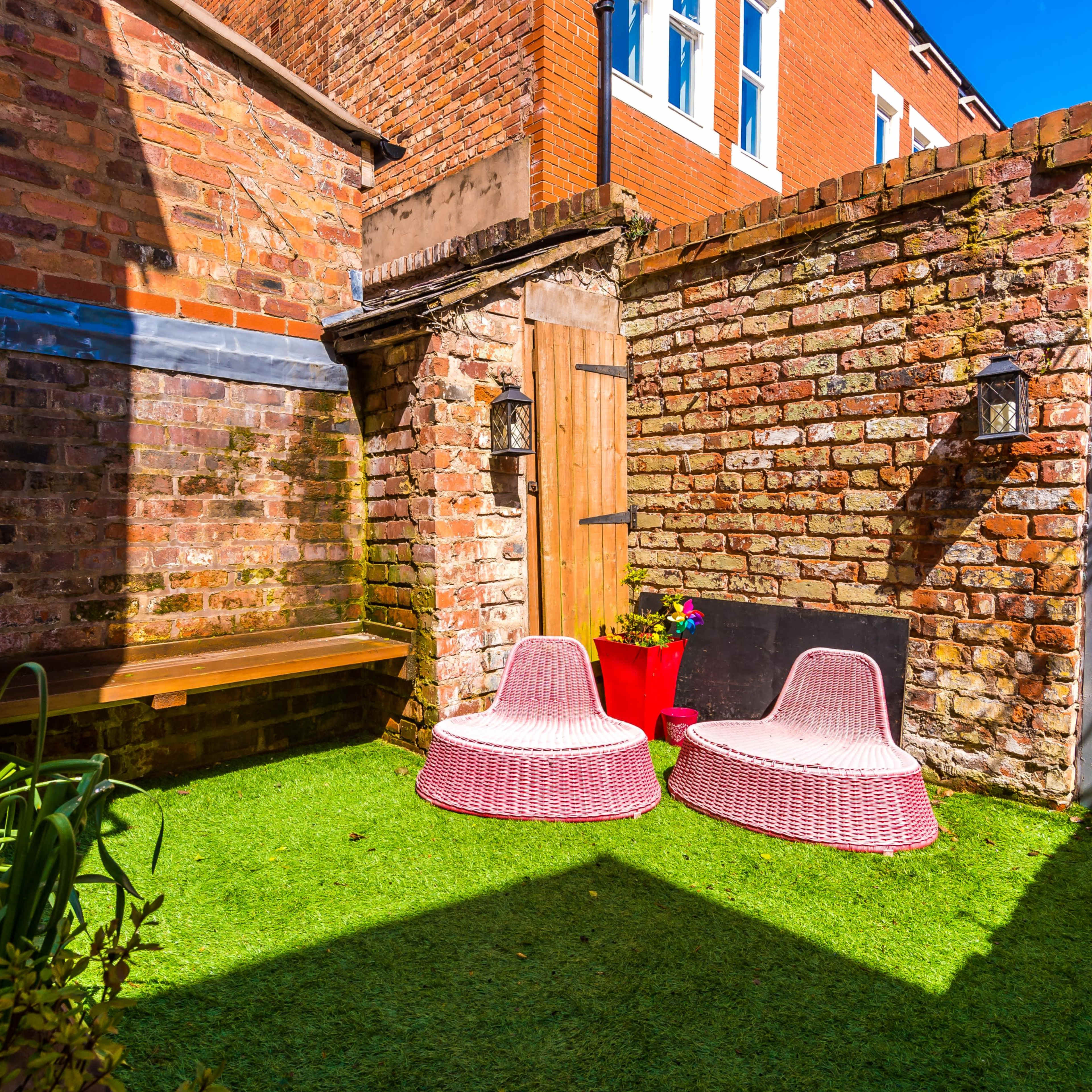 A small courtyard with two pink, wicker chairs on artificial grass, surrounded by brick walls and a wooden door.