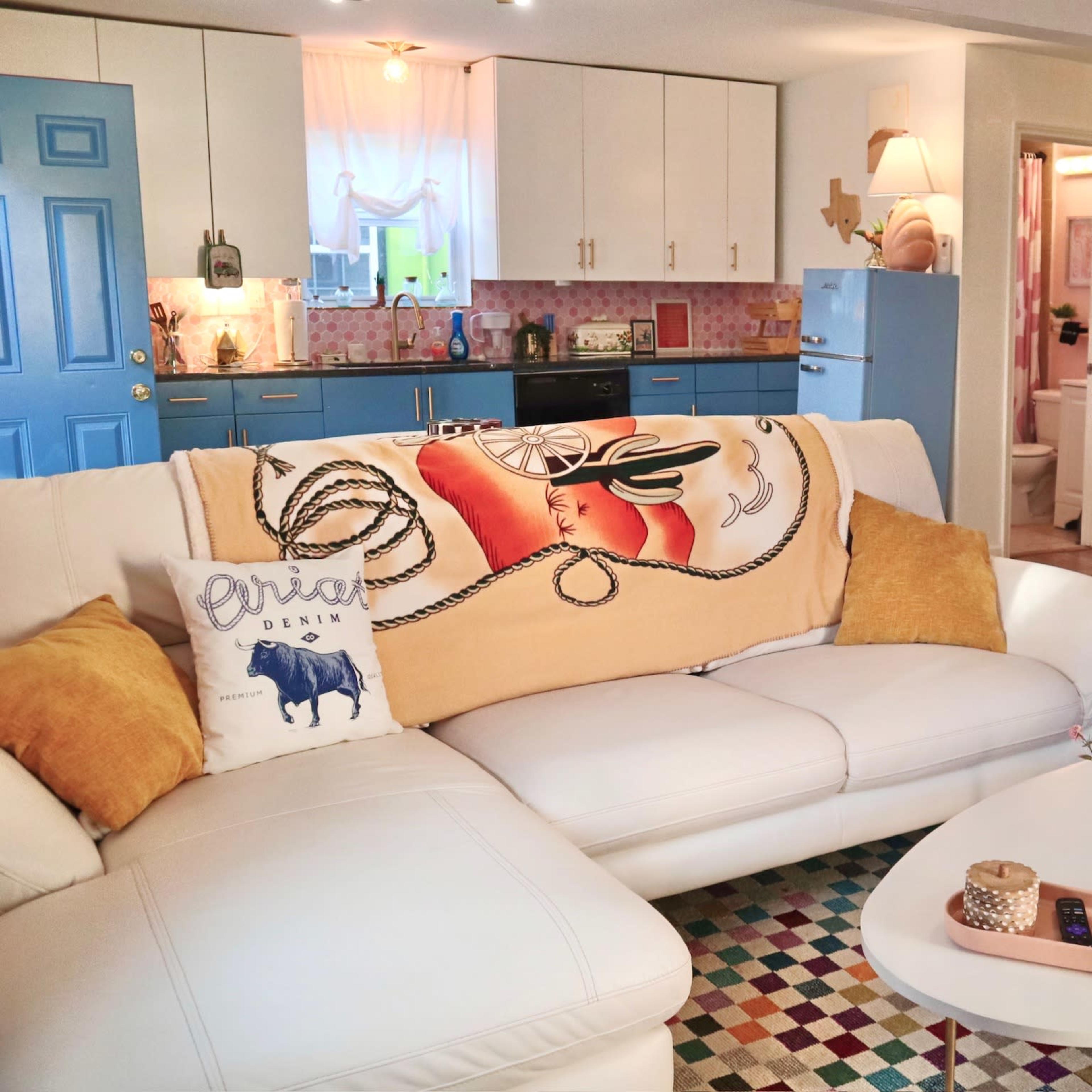 A bright living room features a white sofa adorned with a decorative throw, a colorful area rug, and a kitchen visible in the background with blue cabinets and a vintage refrigerator.