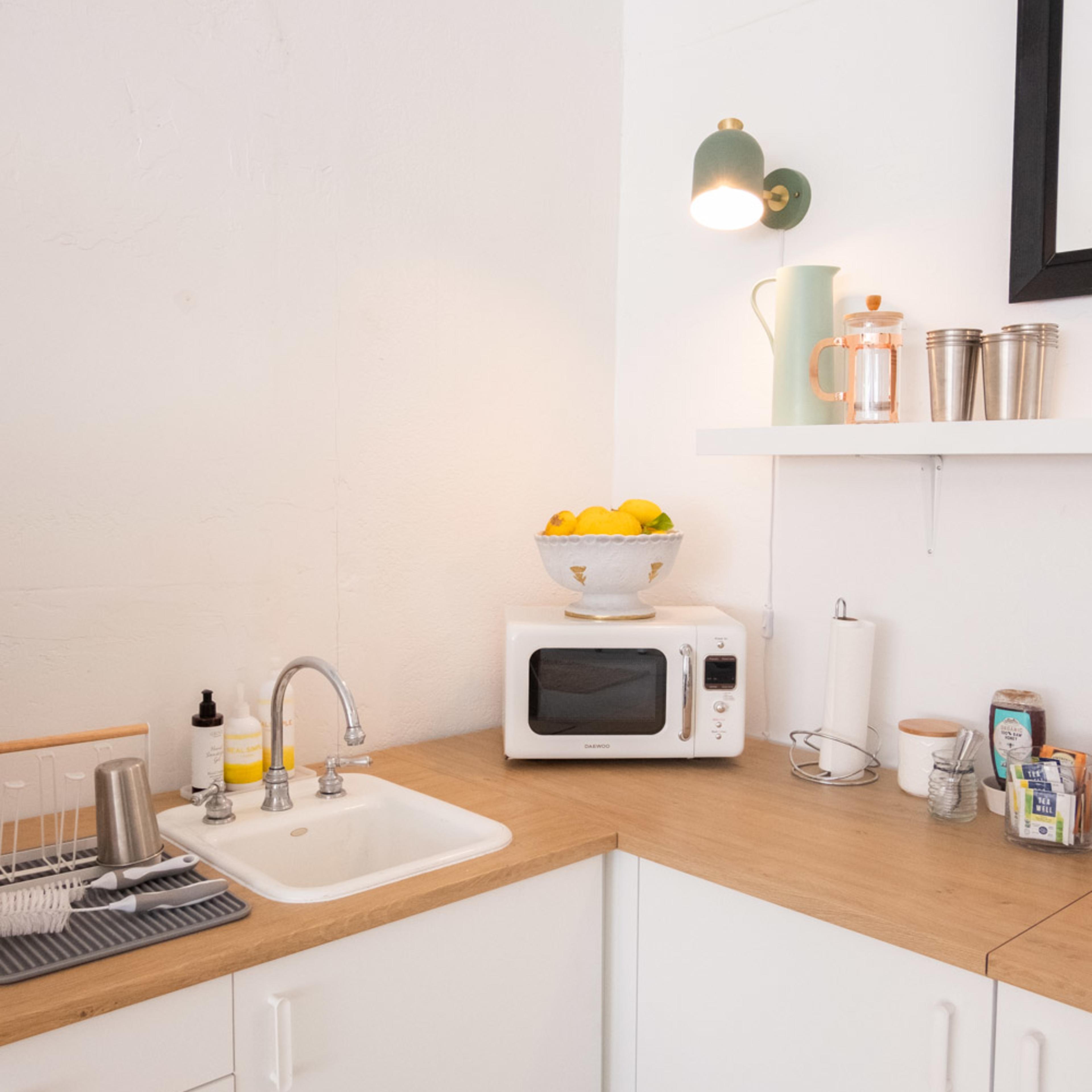 A corner kitchen features a white sink, a microwave on a countertop, a bowl of lemons above, and neatly arranged kitchen items on a shelf.