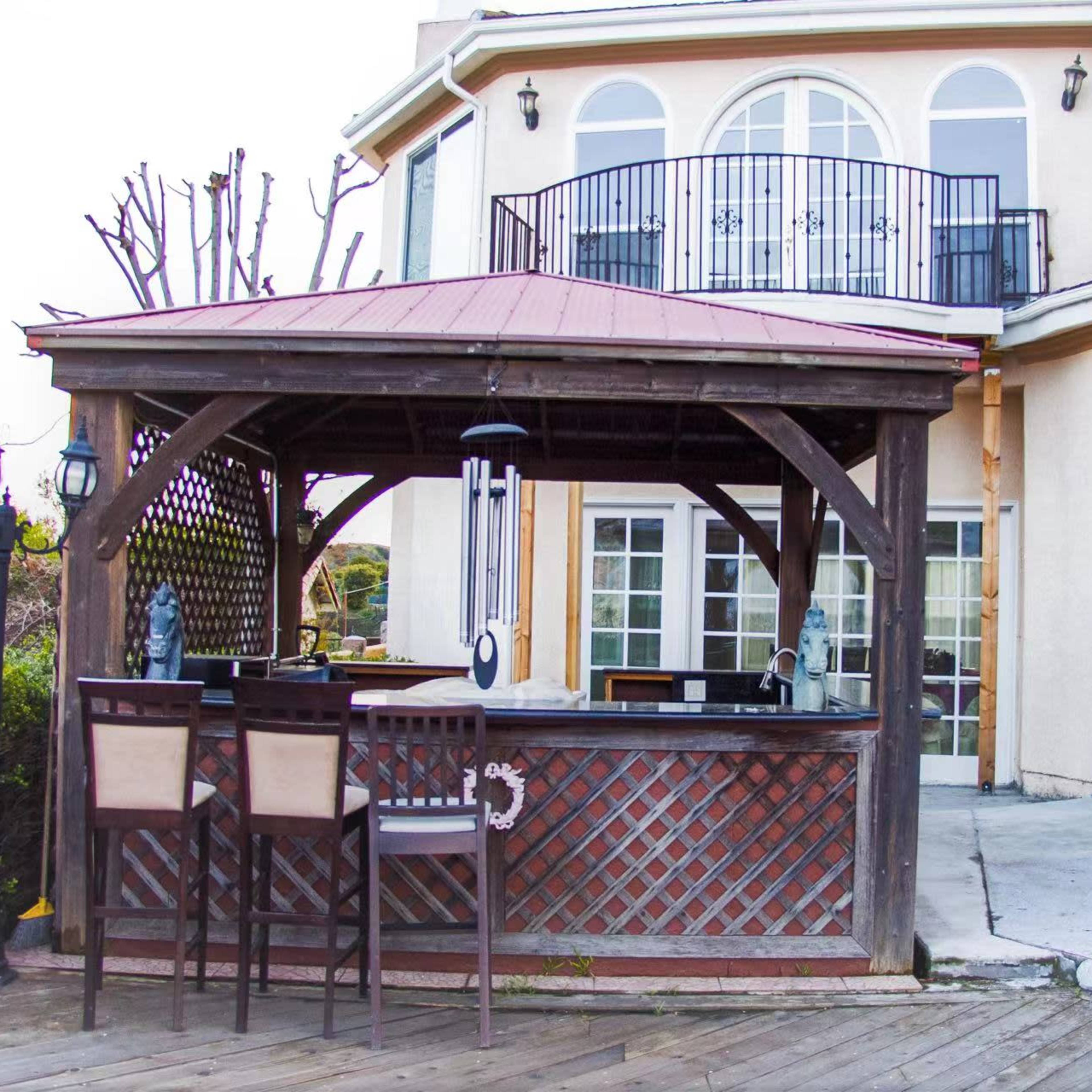 A wooden gazebo-like outdoor bar area with high stools, situated next to a large house with multiple windows and a balcony.