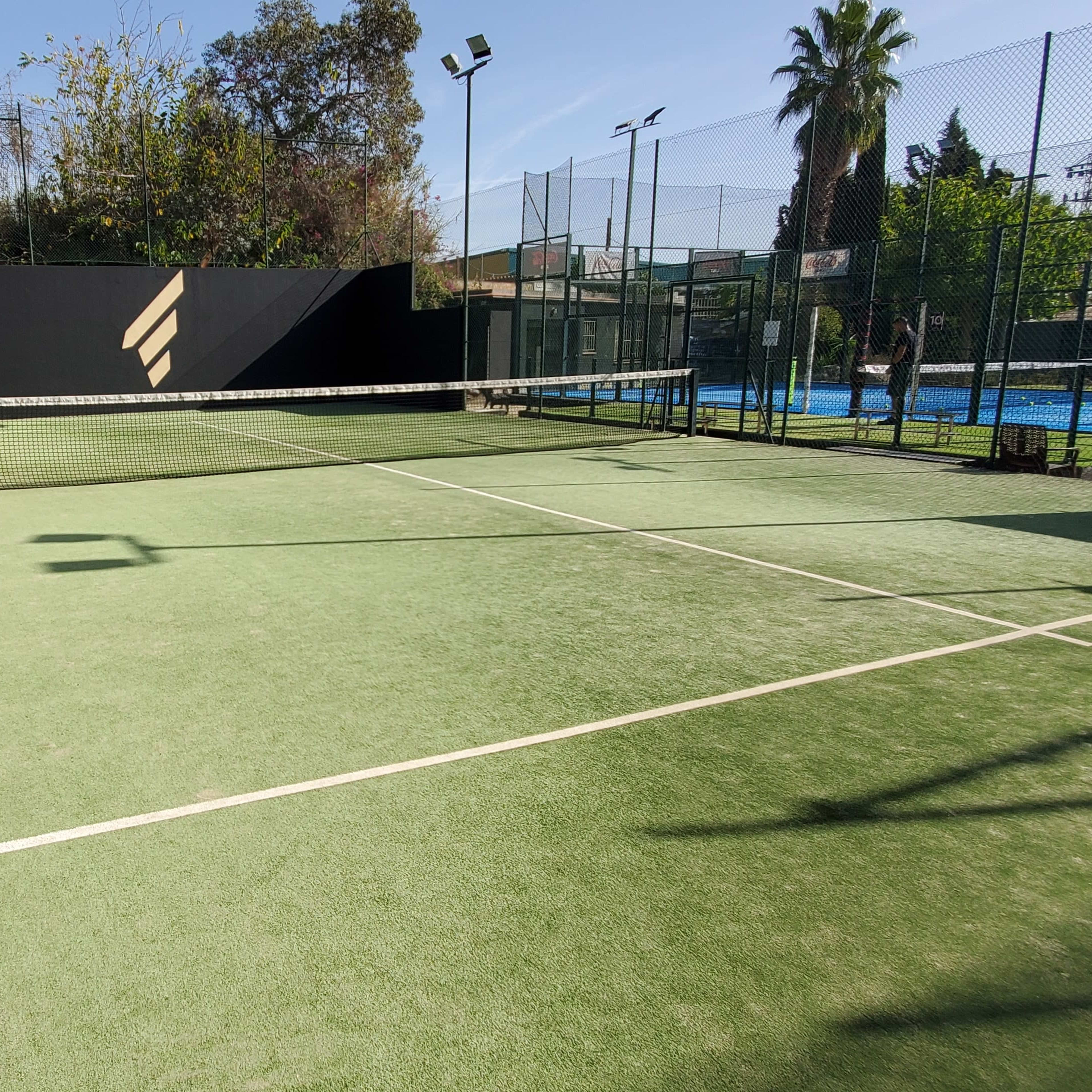 A tennis court with a green surface and a black fence is visible, featuring a net in the center and neighboring fenced courts in the background.