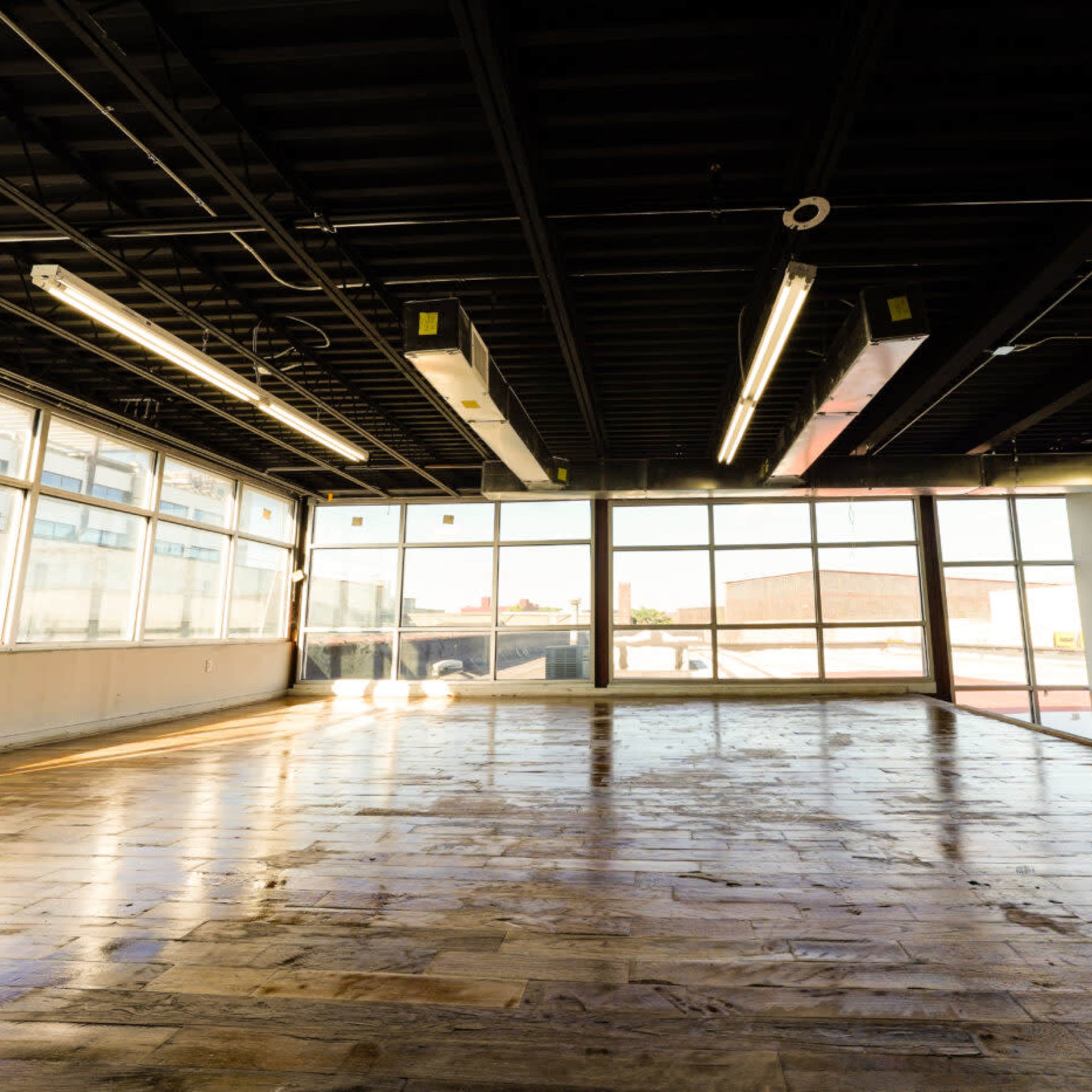 A spacious, empty room with large windows allowing natural light to fill the space and a wooden floor reflecting the light.