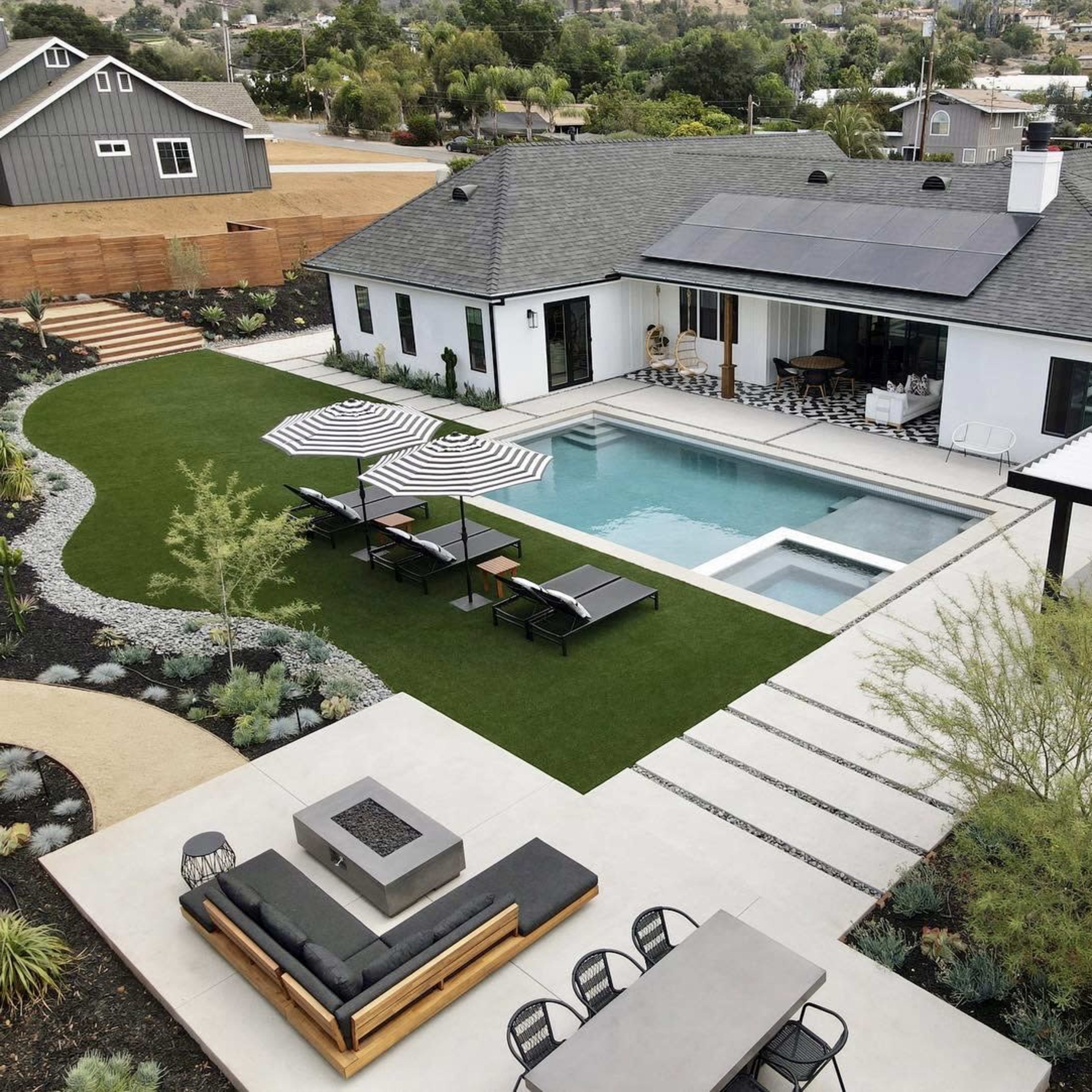 A modern backyard featuring a swimming pool, lounge chairs with umbrellas, a fire pit area, and a landscaped garden surrounding a contemporary house with solar panels on the roof.