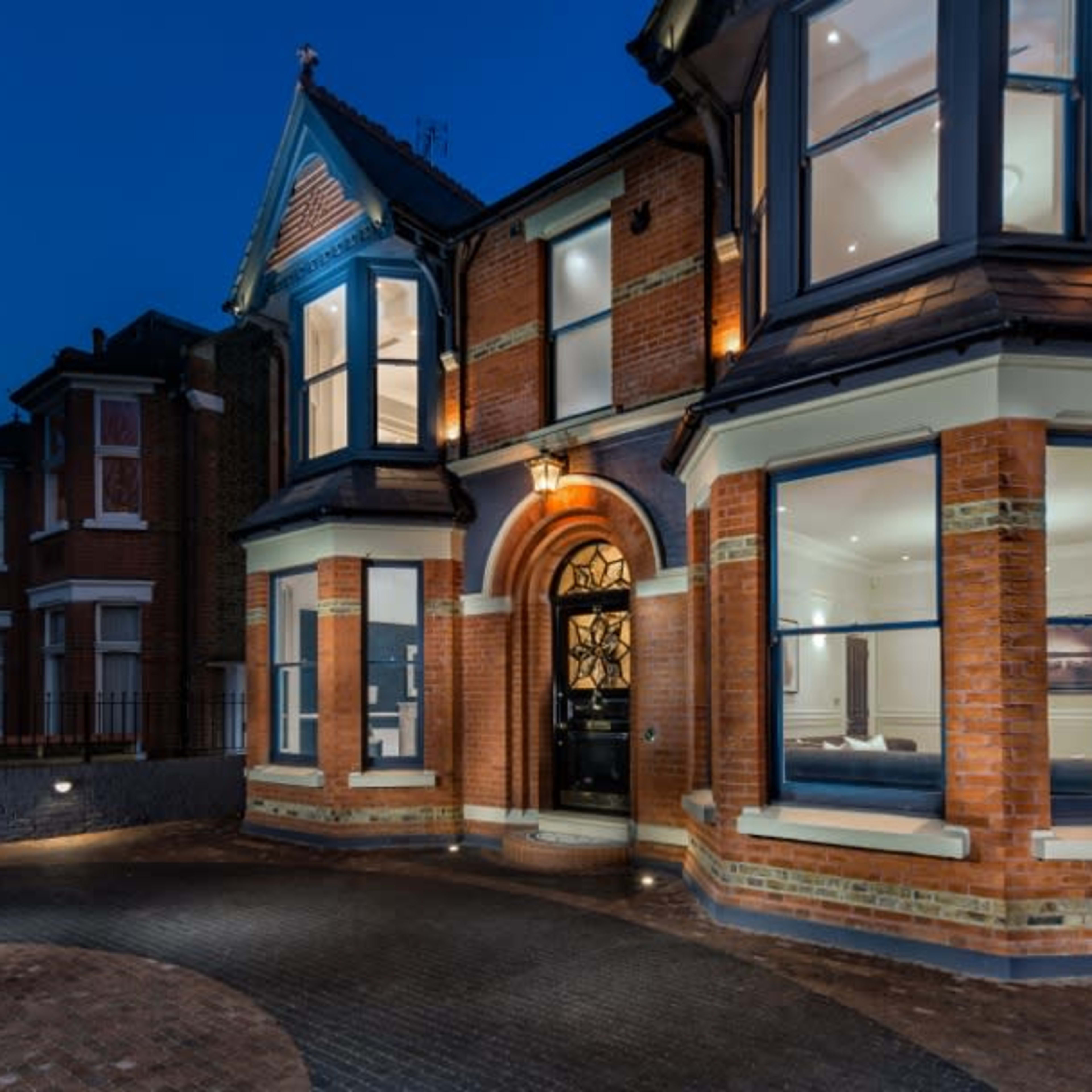 A large, brick-faced house with a prominent entrance and large windows, illuminated against a twilight sky.