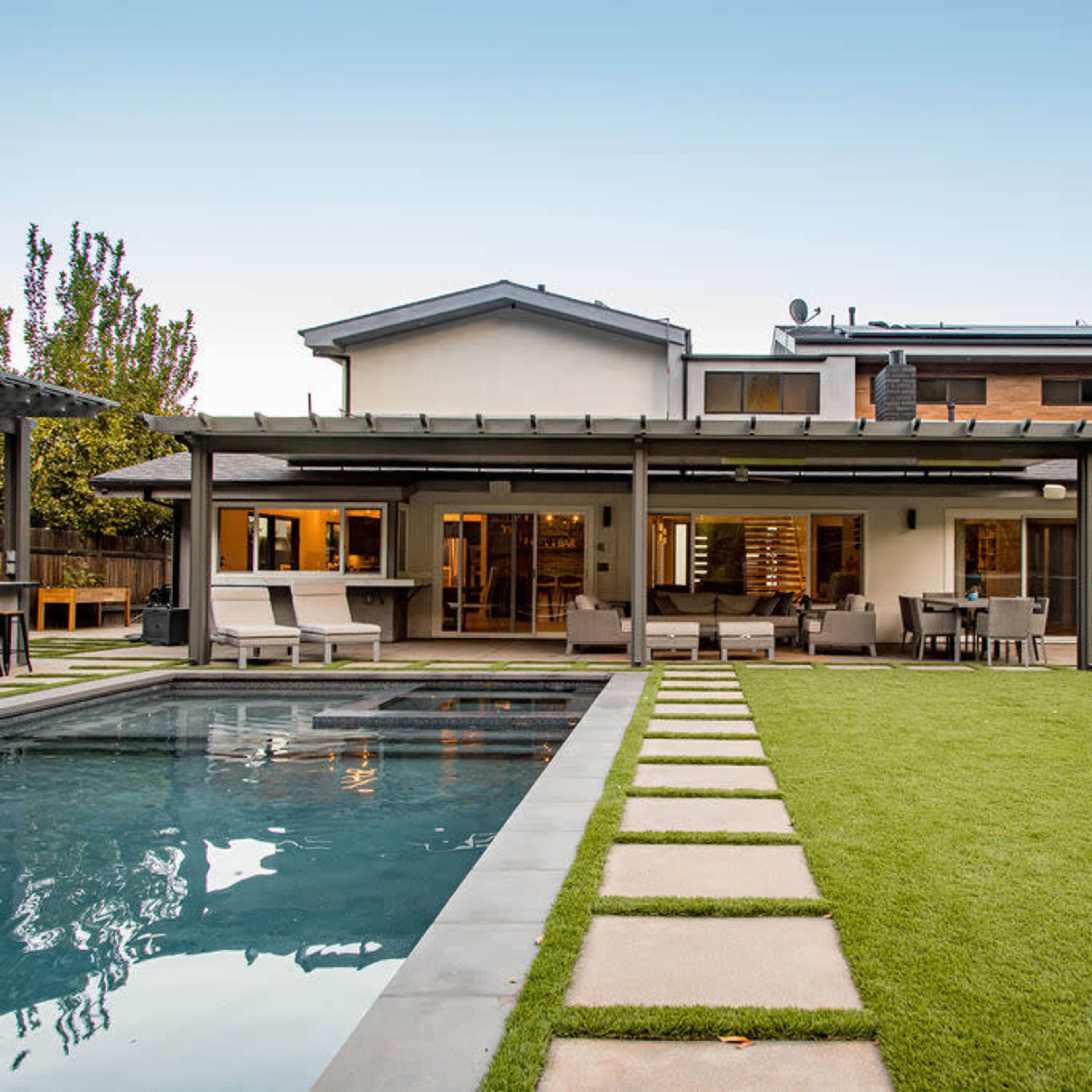 A modern two-story house with a backyard that includes a swimming pool, a patio area with seating, and neatly arranged stone pathways on a lawn.