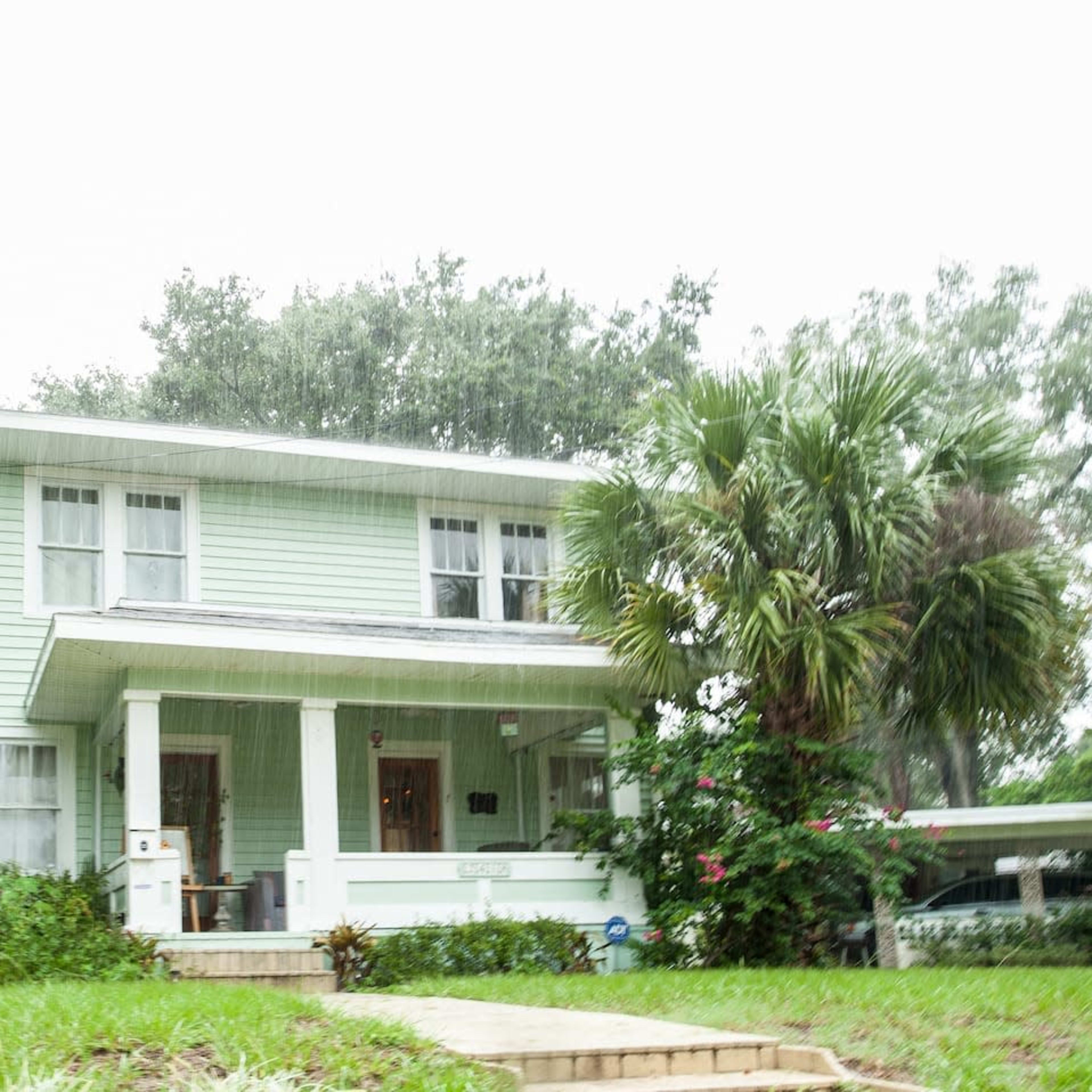 A two-story light green house with white trim and a front porch sits amidst green grass and palm trees in a rainy setting.