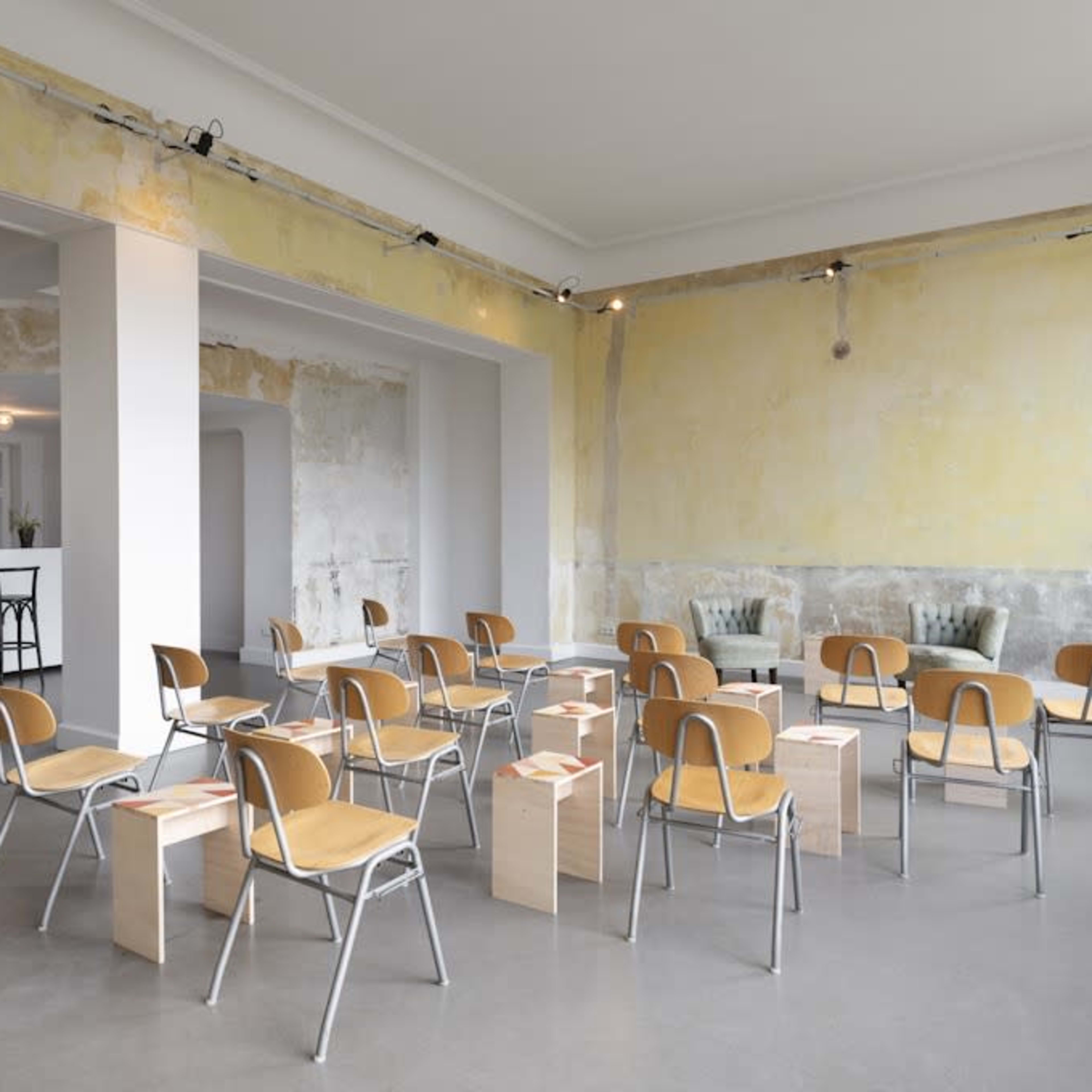 A spacious, minimalist room with several metal and wooden chairs arranged around wooden stools in front of a pale yellow wall.