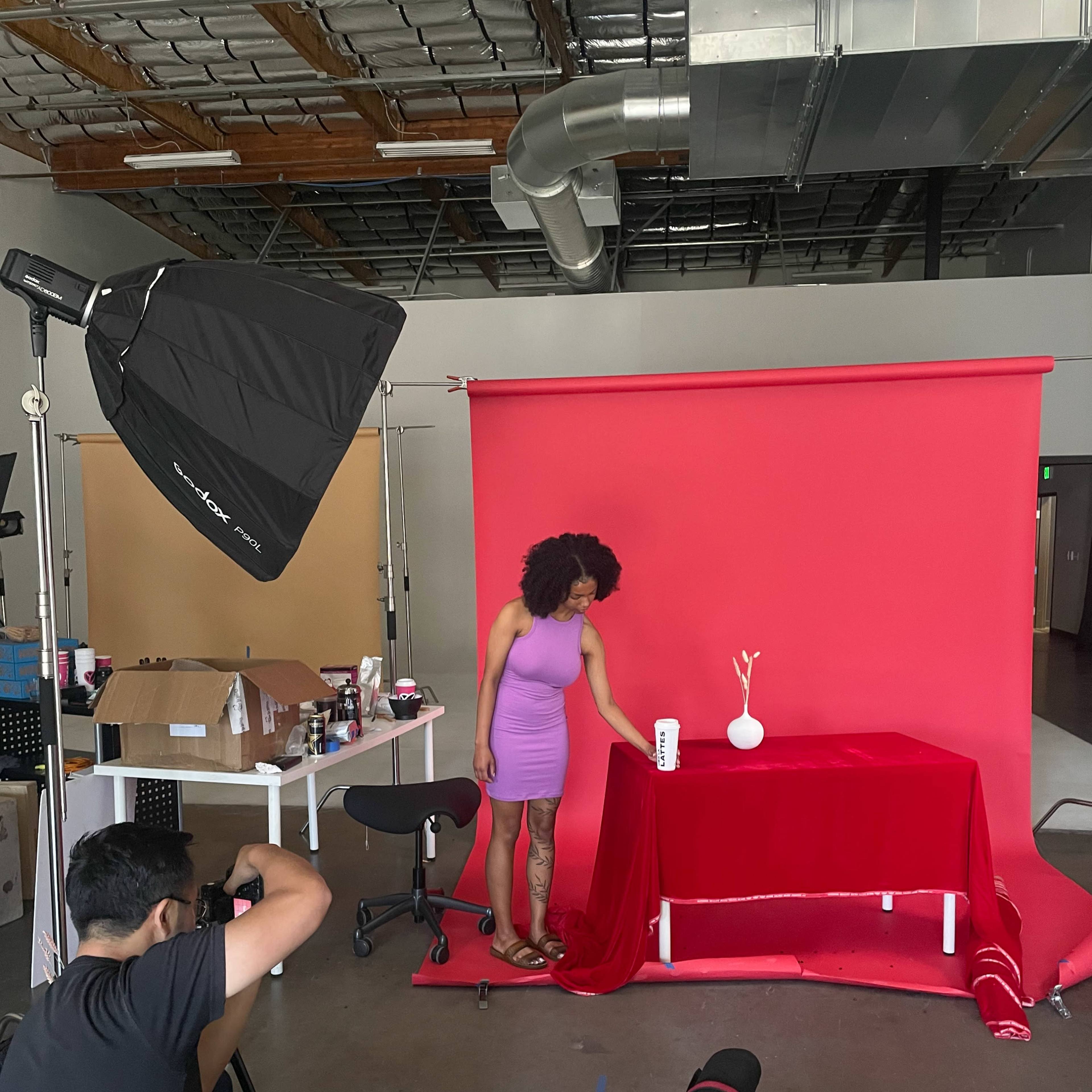 A person adjusts a red tablecloth on a table in front of a red backdrop while another individual takes a photograph in a studio setting.