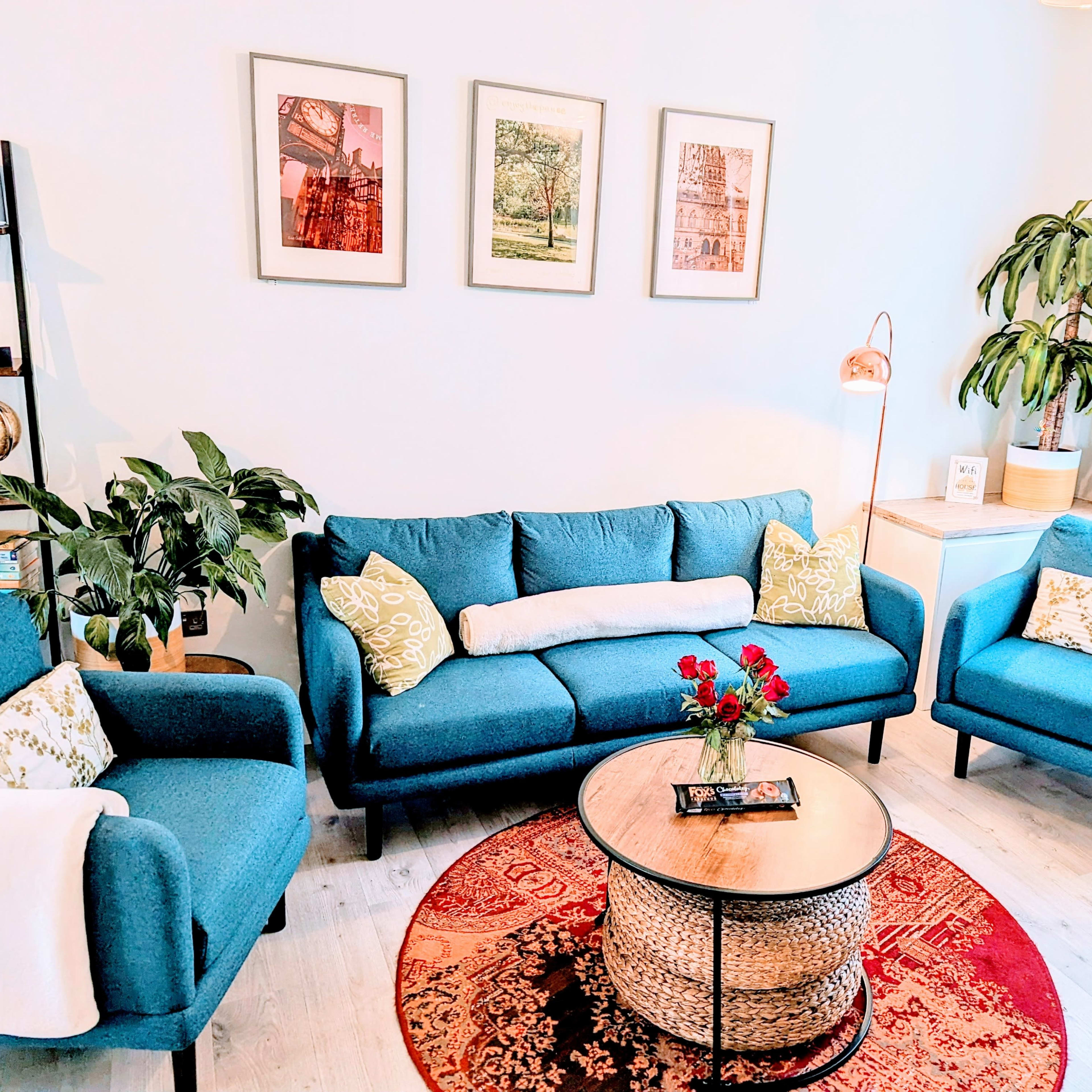 A cozy living room features a blue sofa and armchairs, a round coffee table, a decorative rug, and framed artwork on the walls.