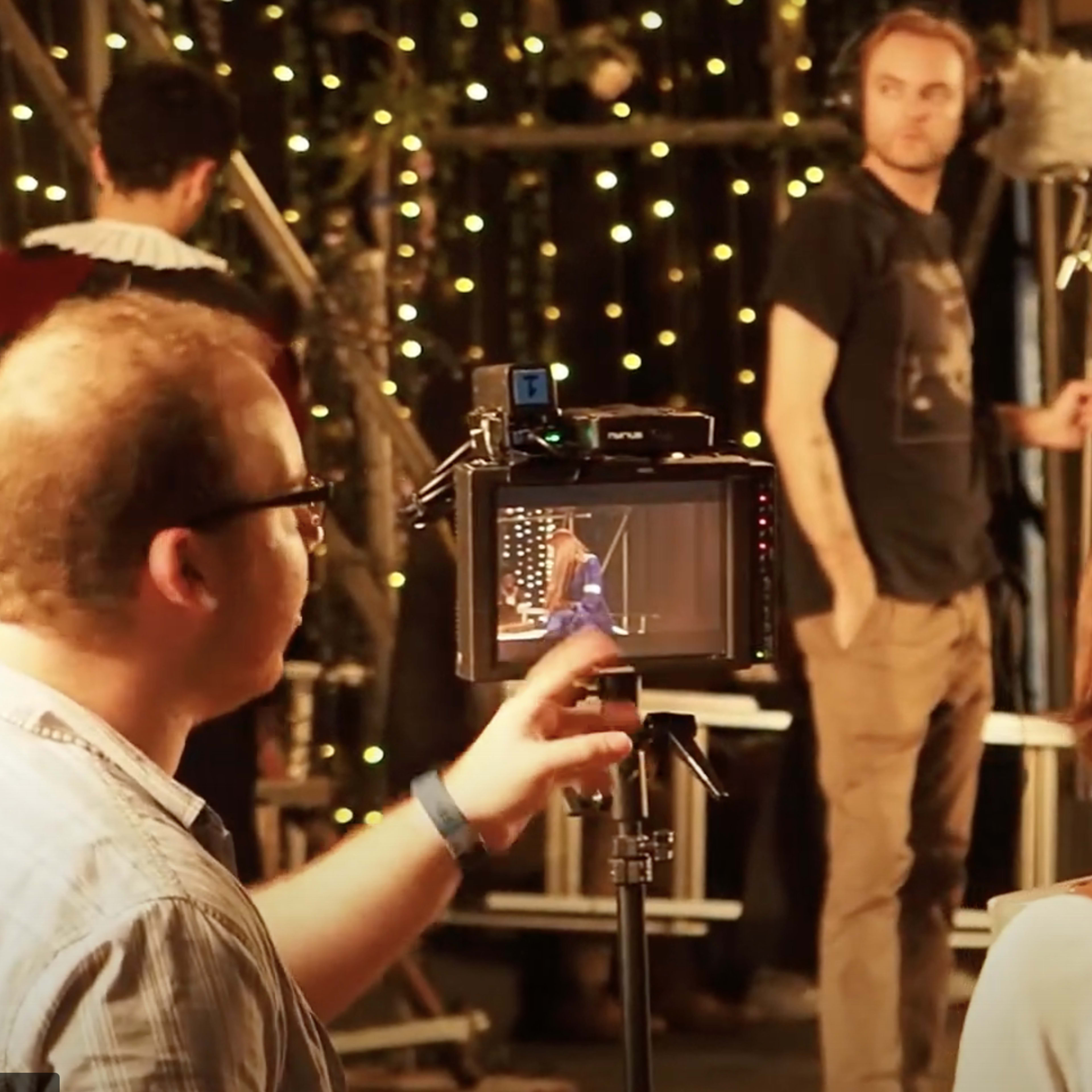 A film crew sets up equipment in a studio with decorative lights while preparing a scene featuring actors in costumes.