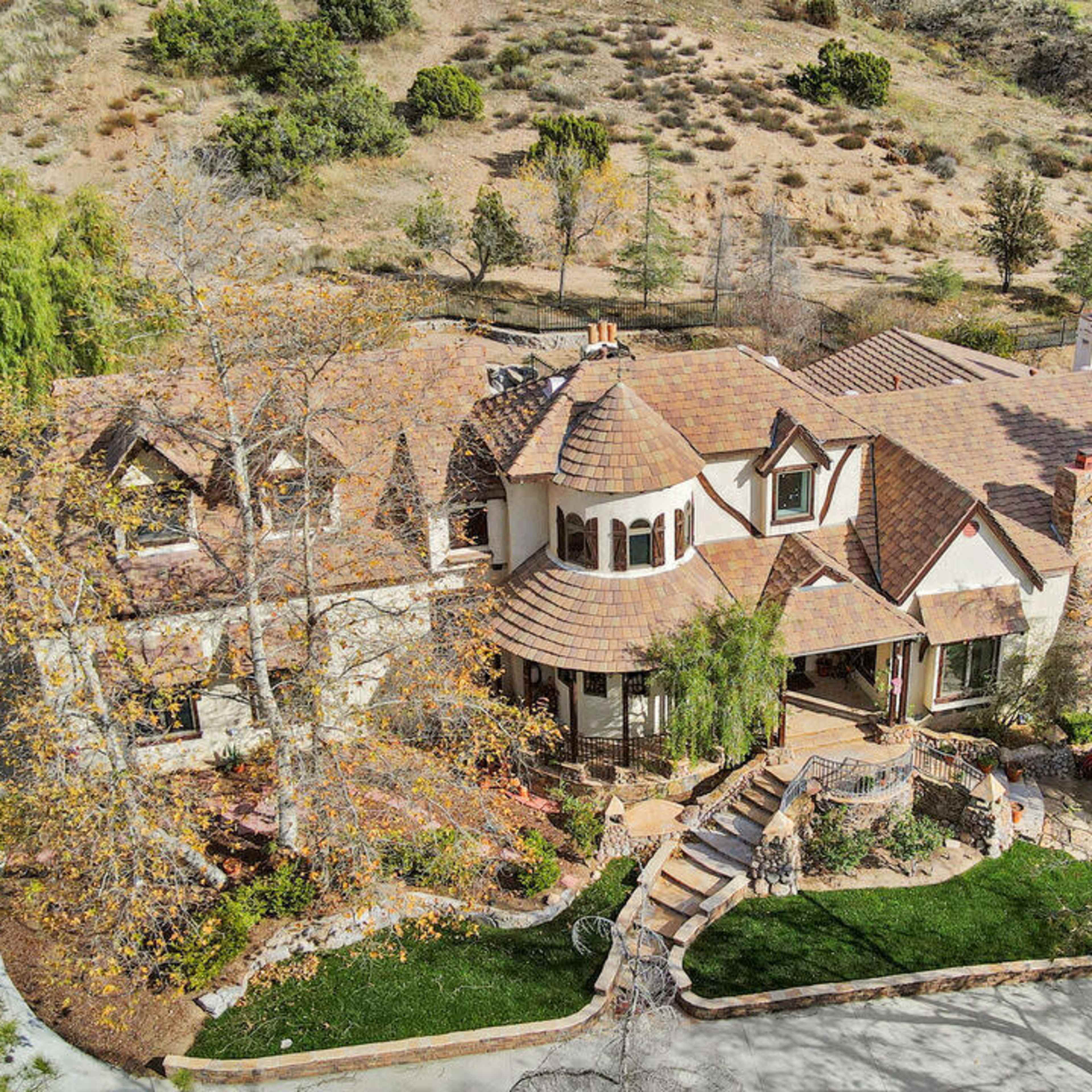 A large, upscale home surrounded by trees and a landscaped yard, with winding pathways and a hilly backdrop.