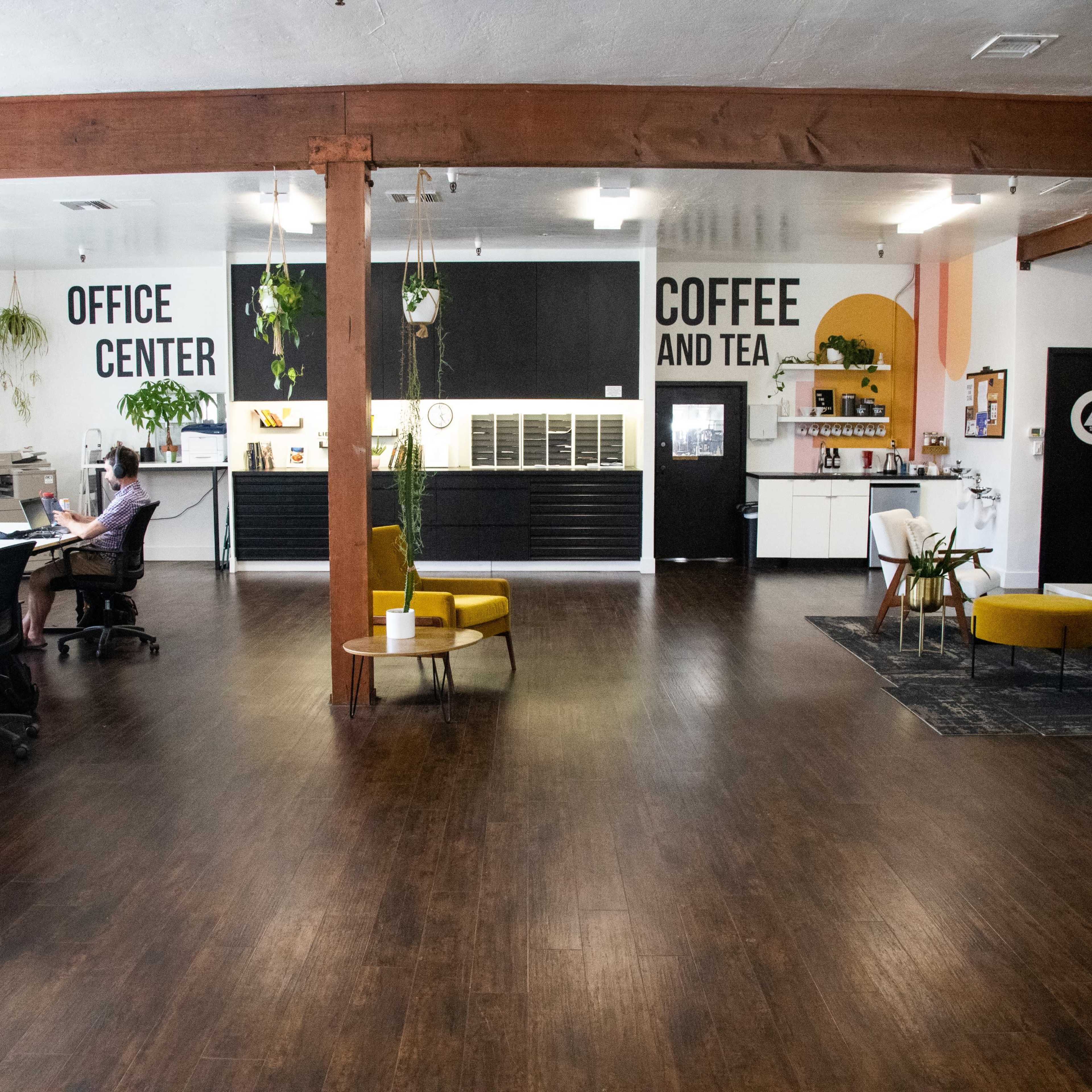 A modern office space featuring workstations, a coffee area, and ample greenery, with a prominent sign that reads "OFFICE CENTER" and "COFFEE AND TEA."