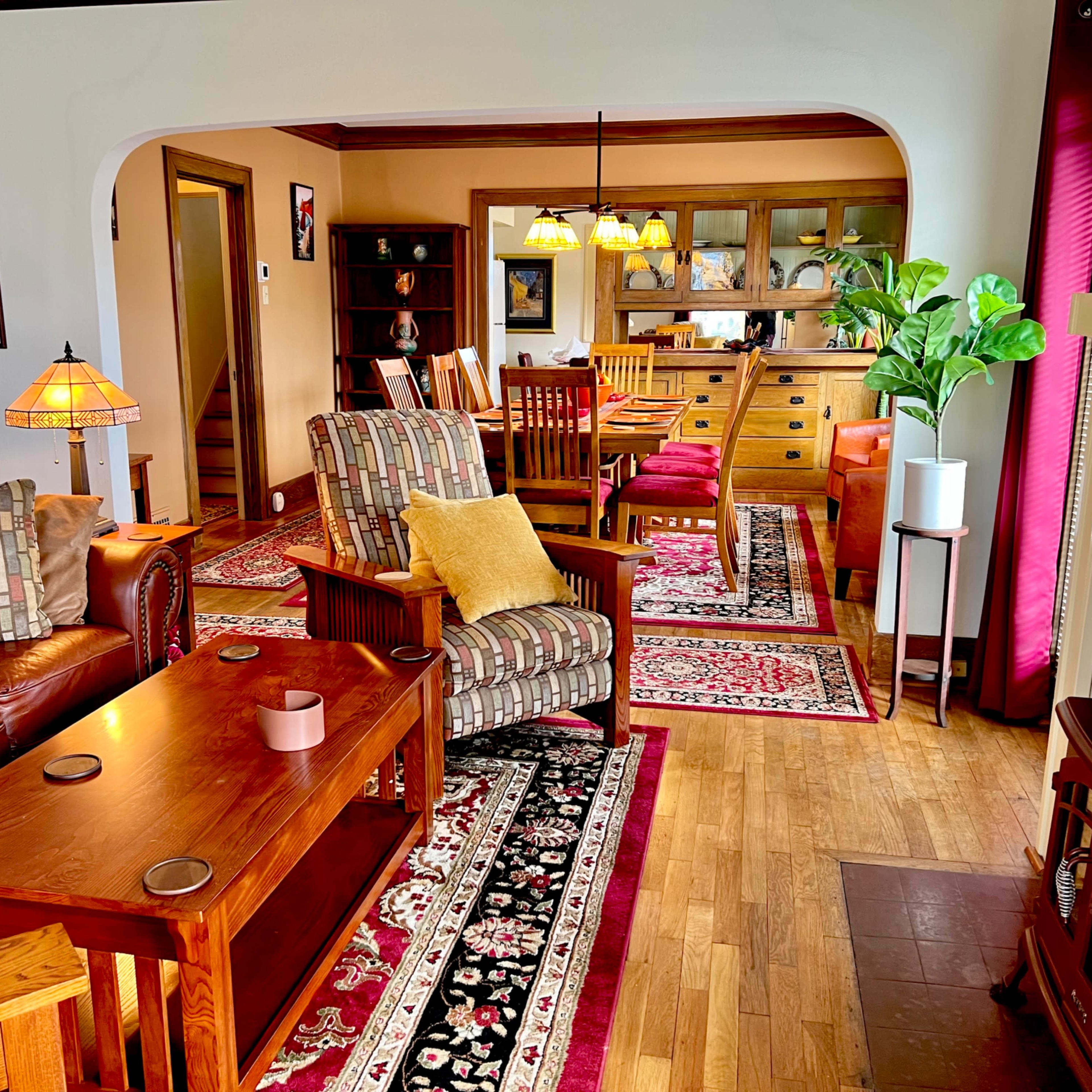 A cozy living room with a mix of wooden furniture, patterned rugs, and a fireplace, connected to a dining area visible in the background.