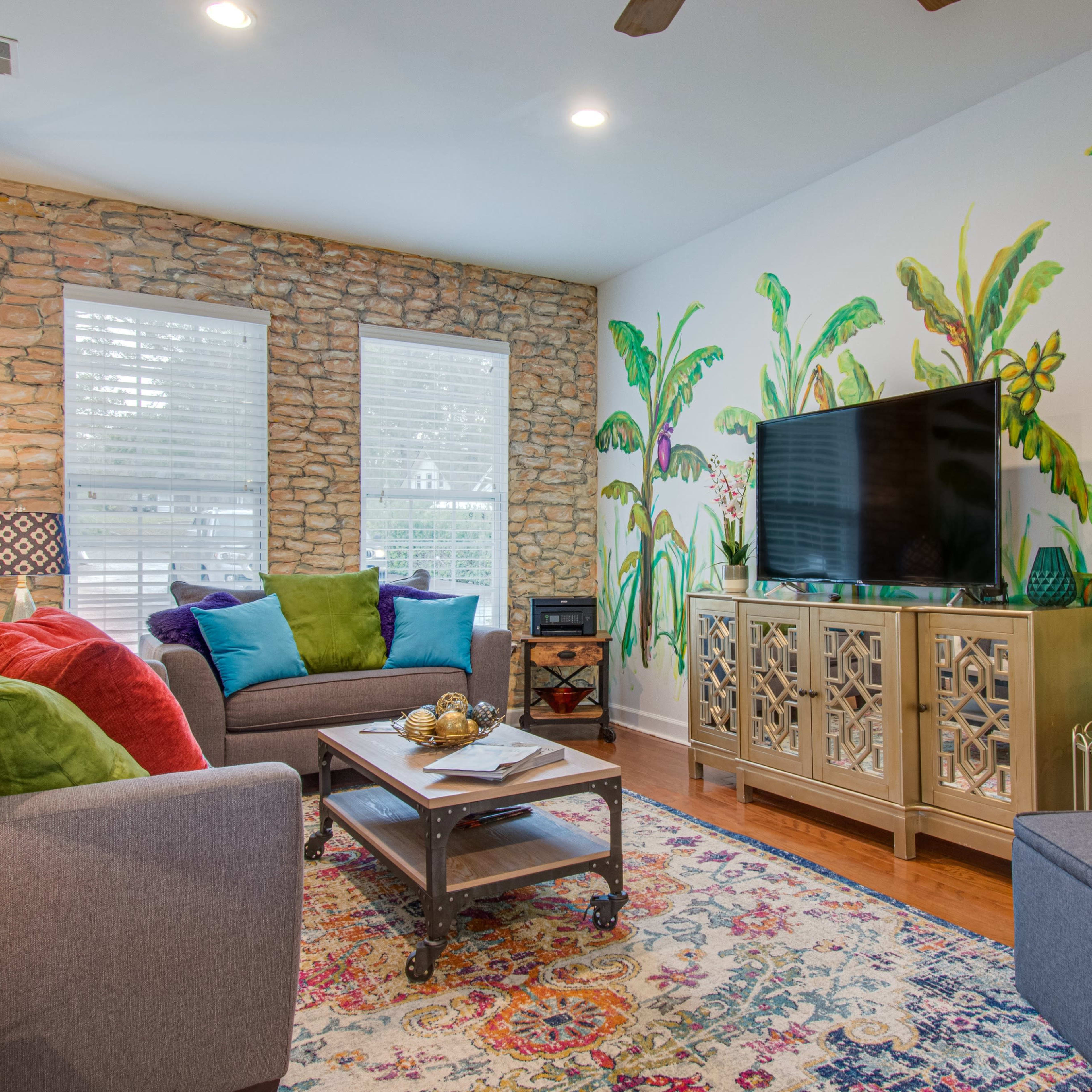 A living room features a stone accent wall, colorful sofa cushions, a wooden coffee table, and a television against a backdrop of tropical wall murals.