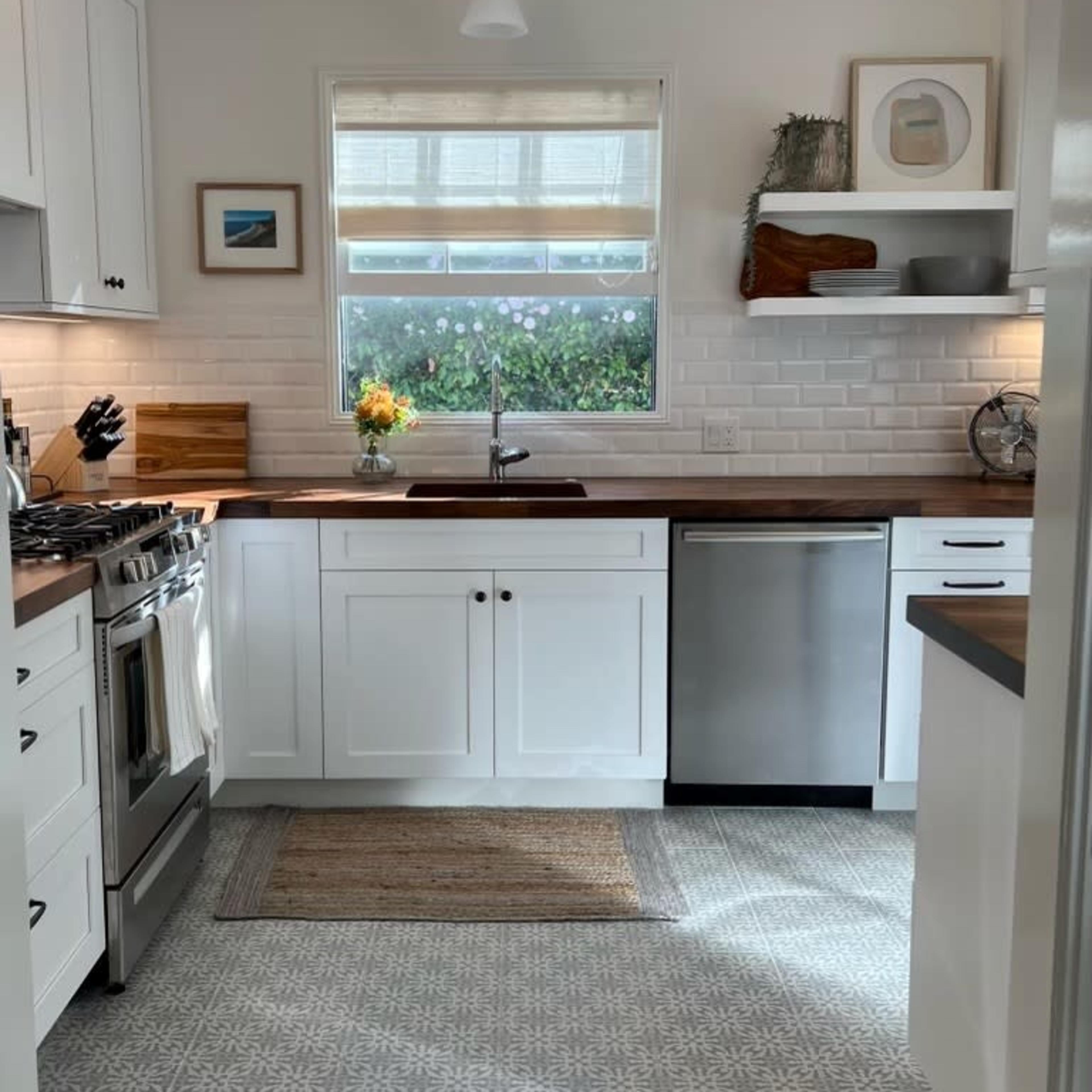 A modern kitchen features white cabinetry, a stainless steel refrigerator, a large window above the sink, and a patterned floor.