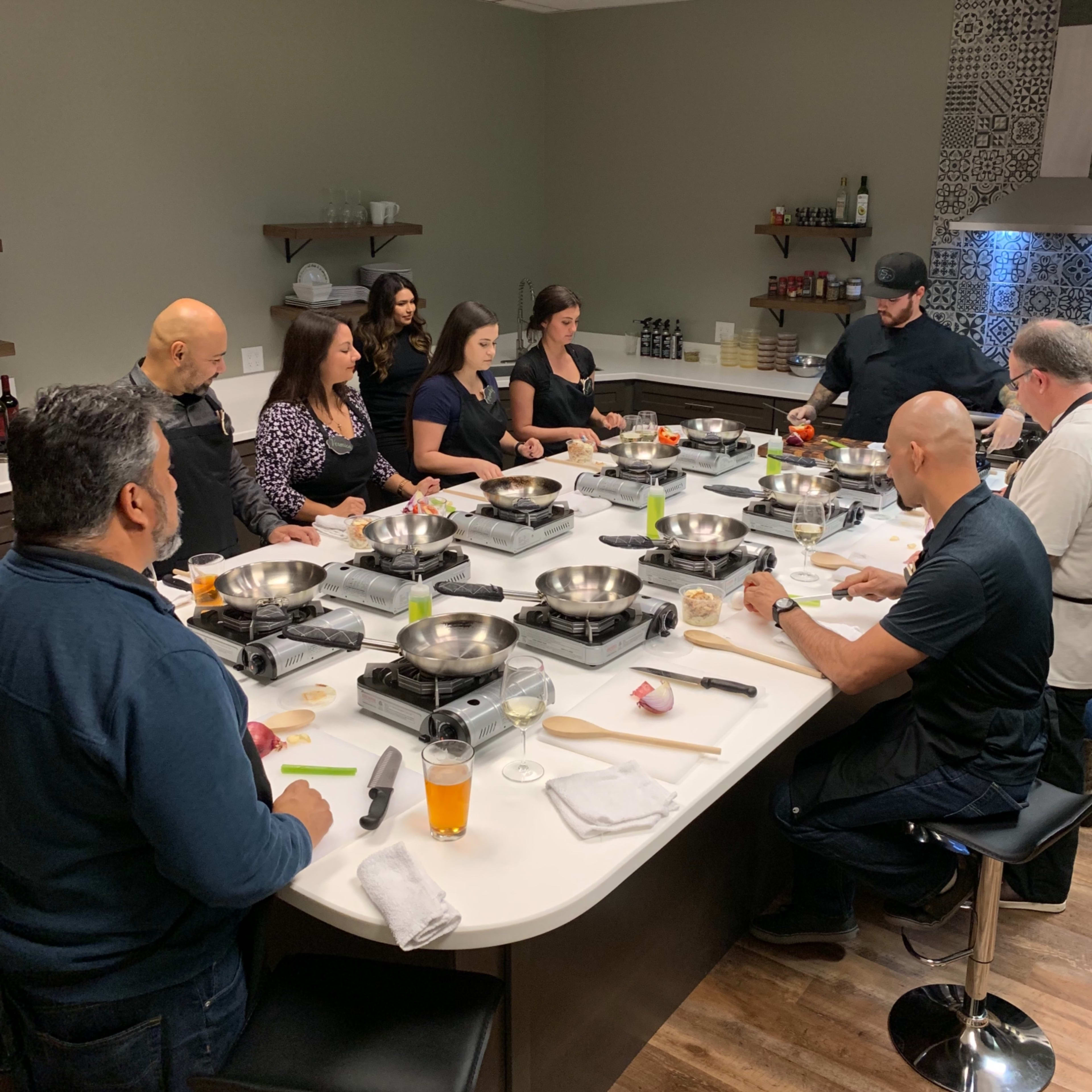 A group of people is gathered around a kitchen island, preparing food using various cooking tools and ingredients.
