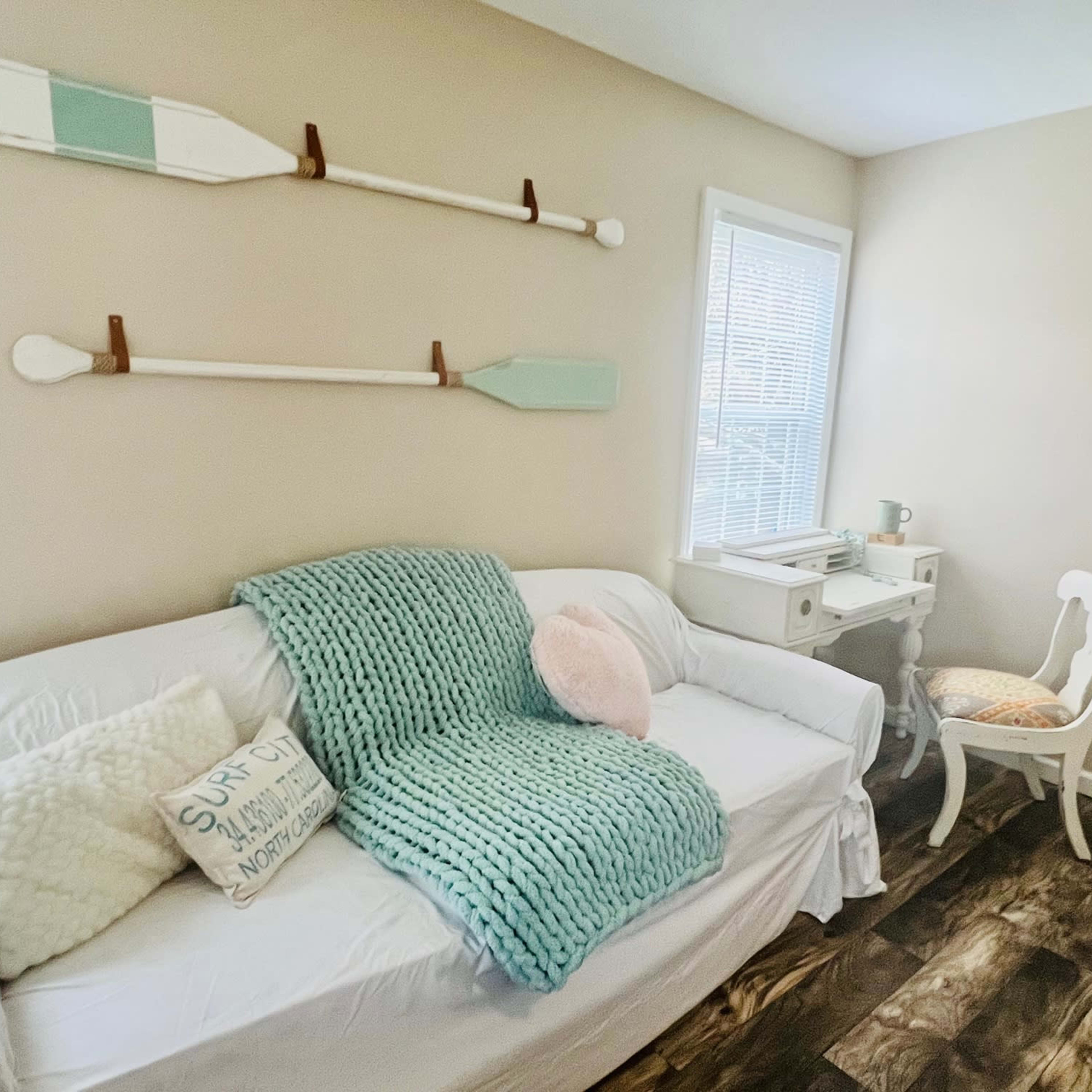 A cozy room featuring a white couch with a blue blanket, decorative pillows, and a small desk next to a chair.