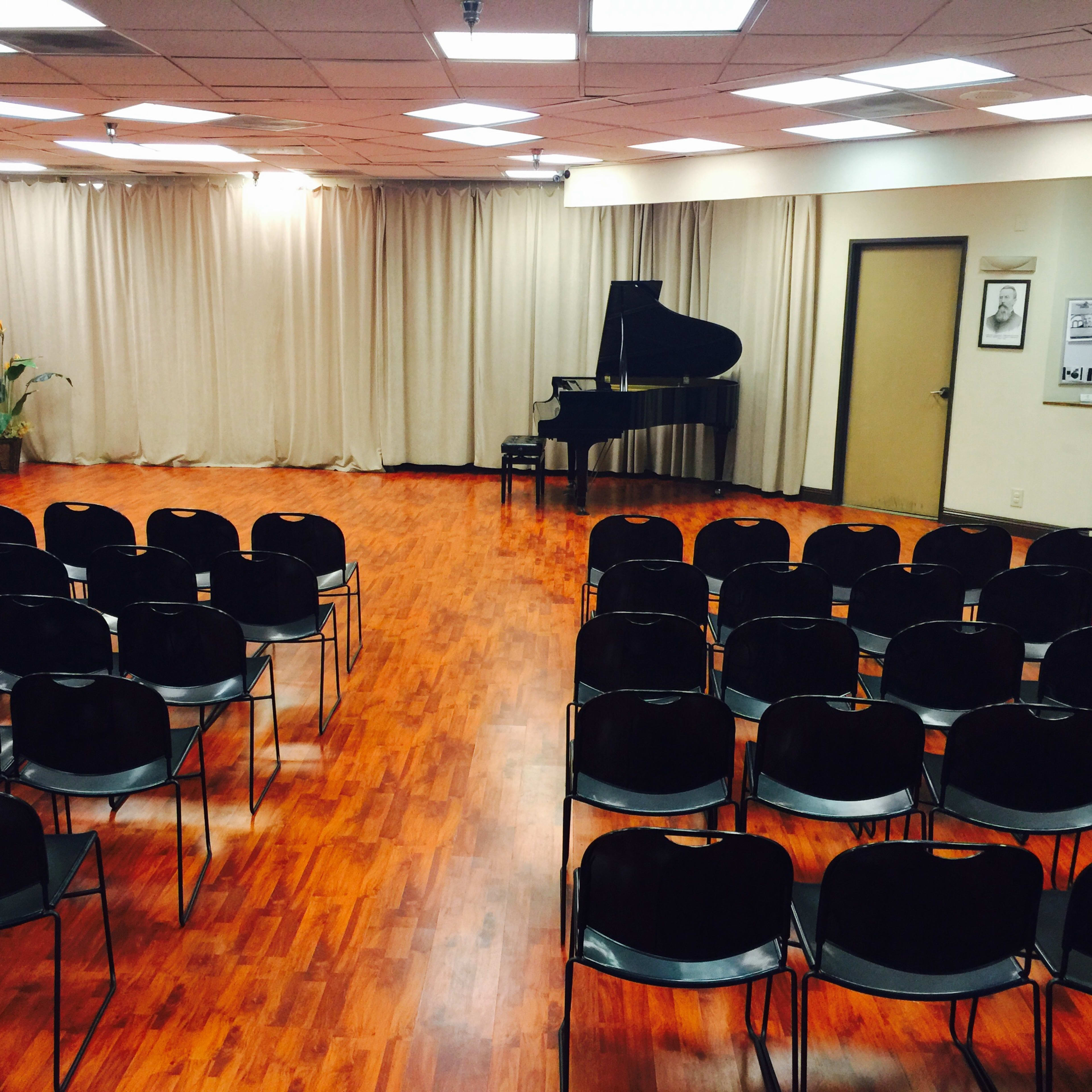 A music performance room with rows of black chairs facing a grand piano and a curtain backdrop.