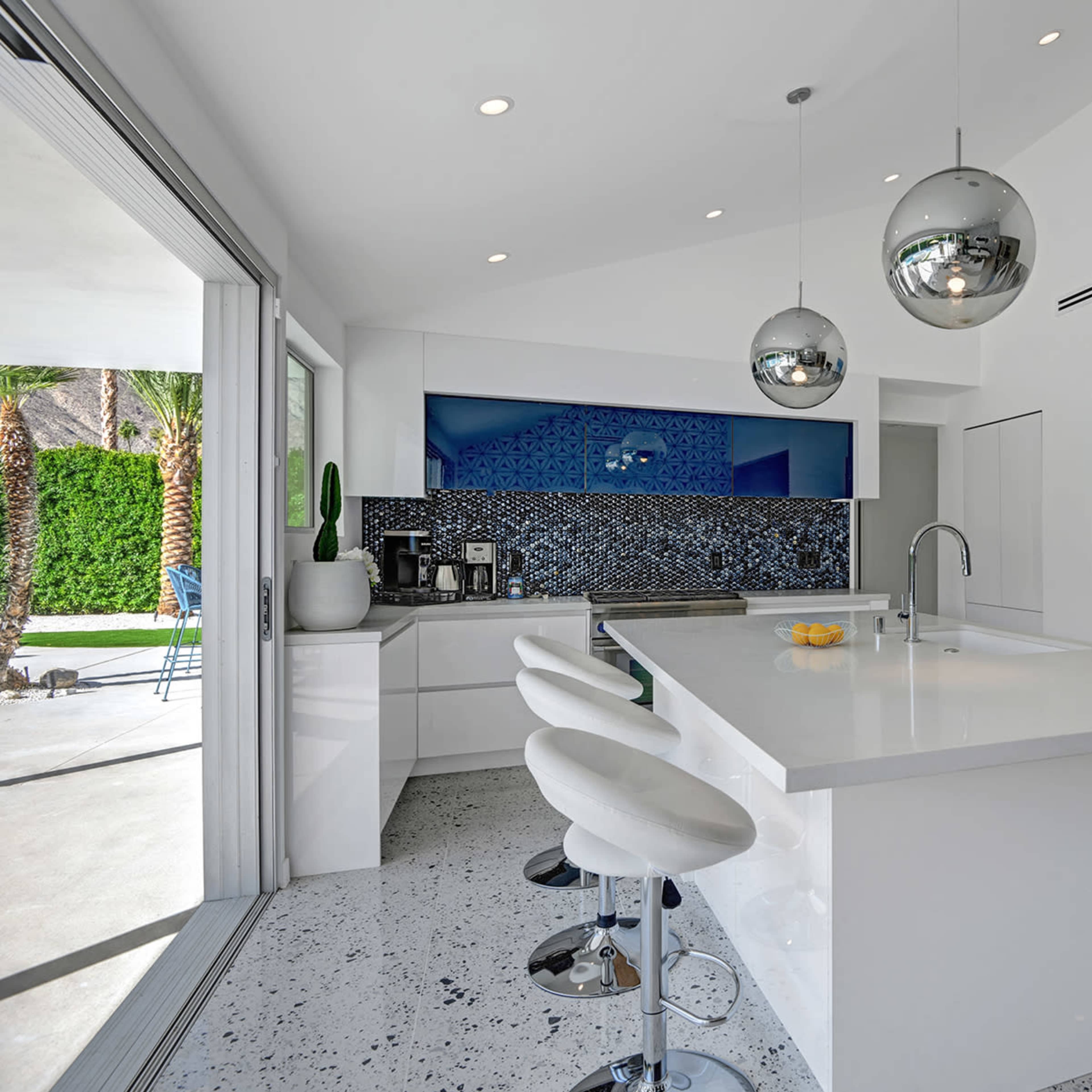 A modern kitchen features a large island with white countertops, three white bar stools, and sleek pendant lights, set against blue mosaic tile and large windows overlooking a patio.