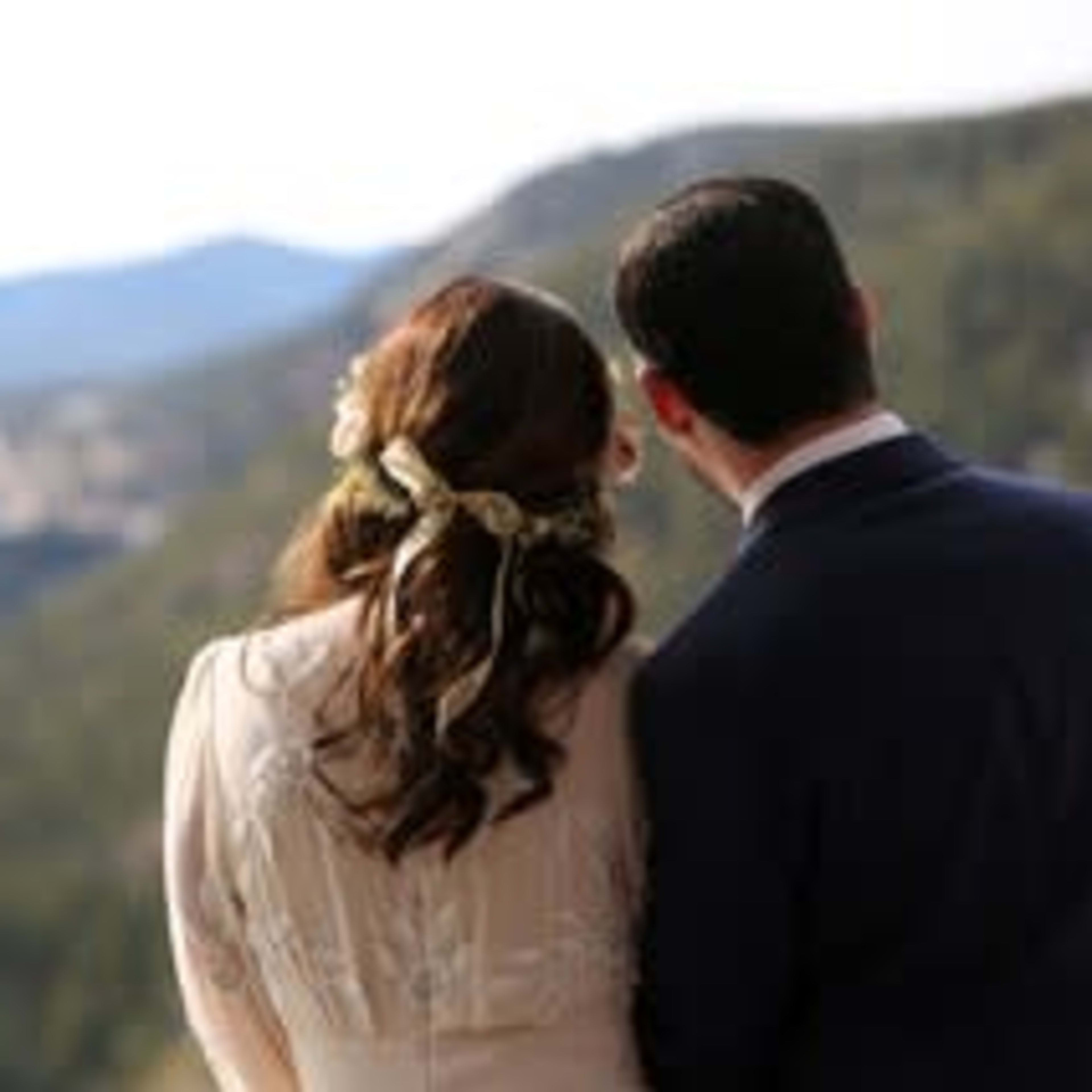 A couple stands together, overlooking a mountain landscape, with the bride's hair adorned with flowers.