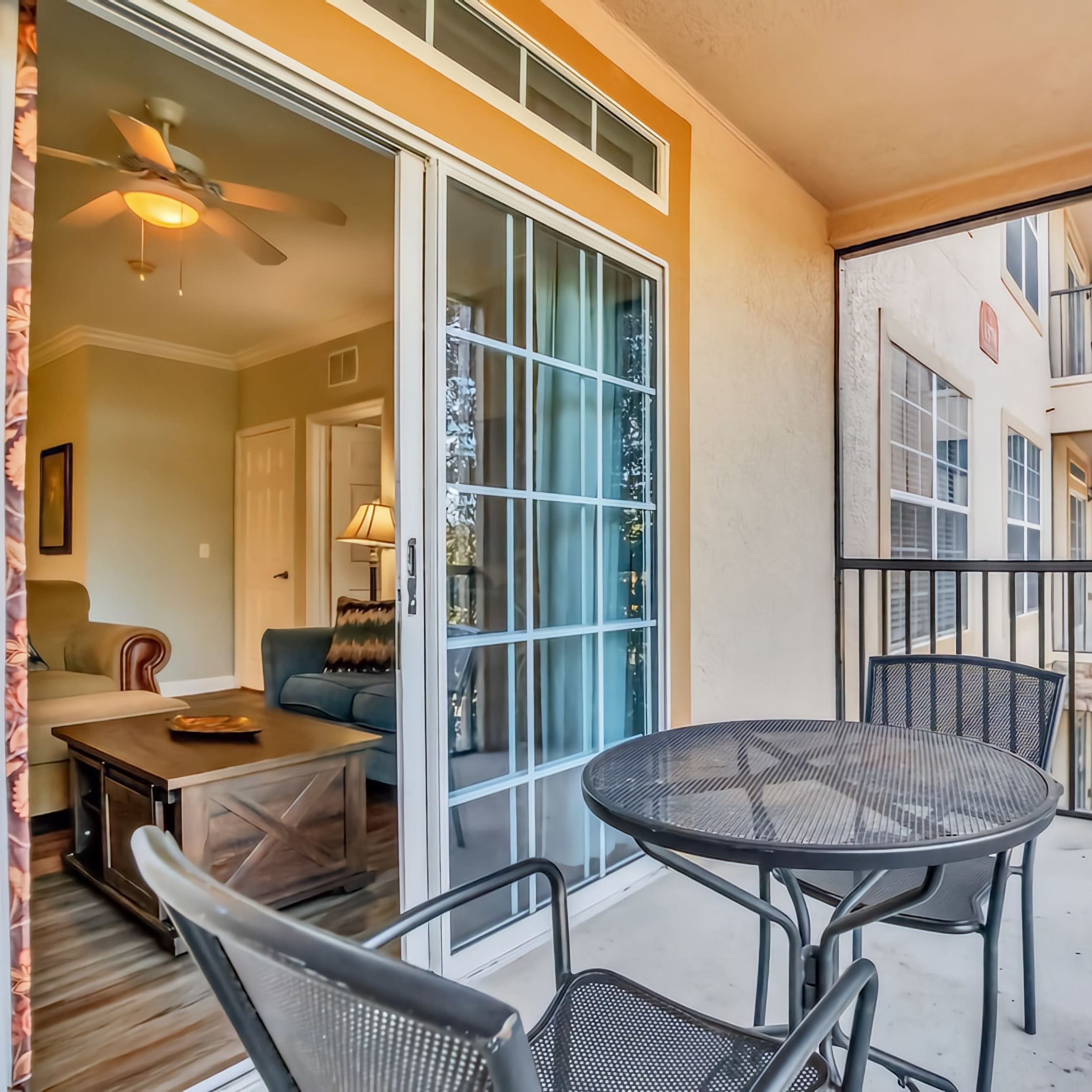 A small balcony features a round table with two chairs, overlooking a view of a nearby building and bright natural light.