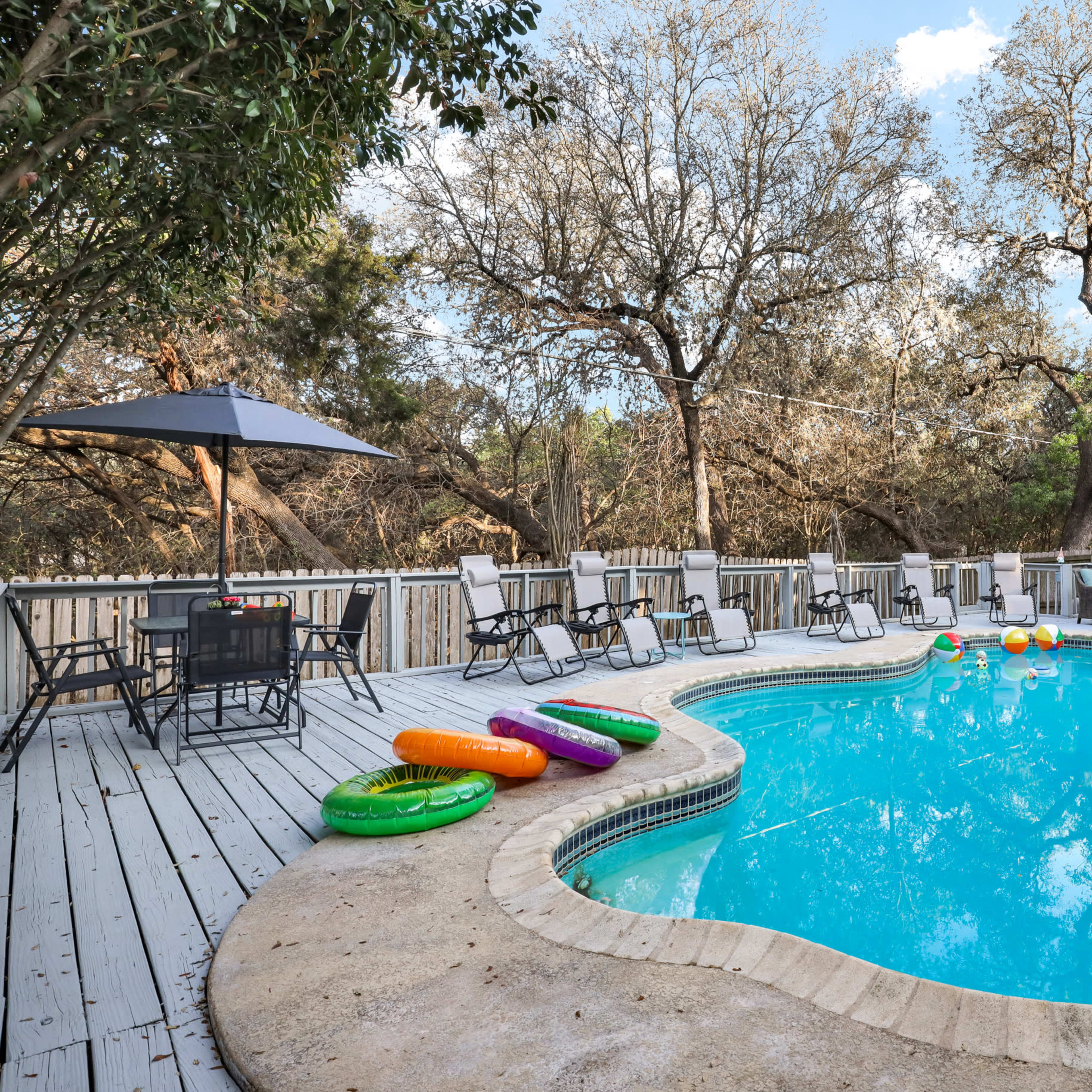 A swimming pool with colorful floats is surrounded by a wooden deck and lawn chairs under an umbrella, situated in a wooded outdoor area.