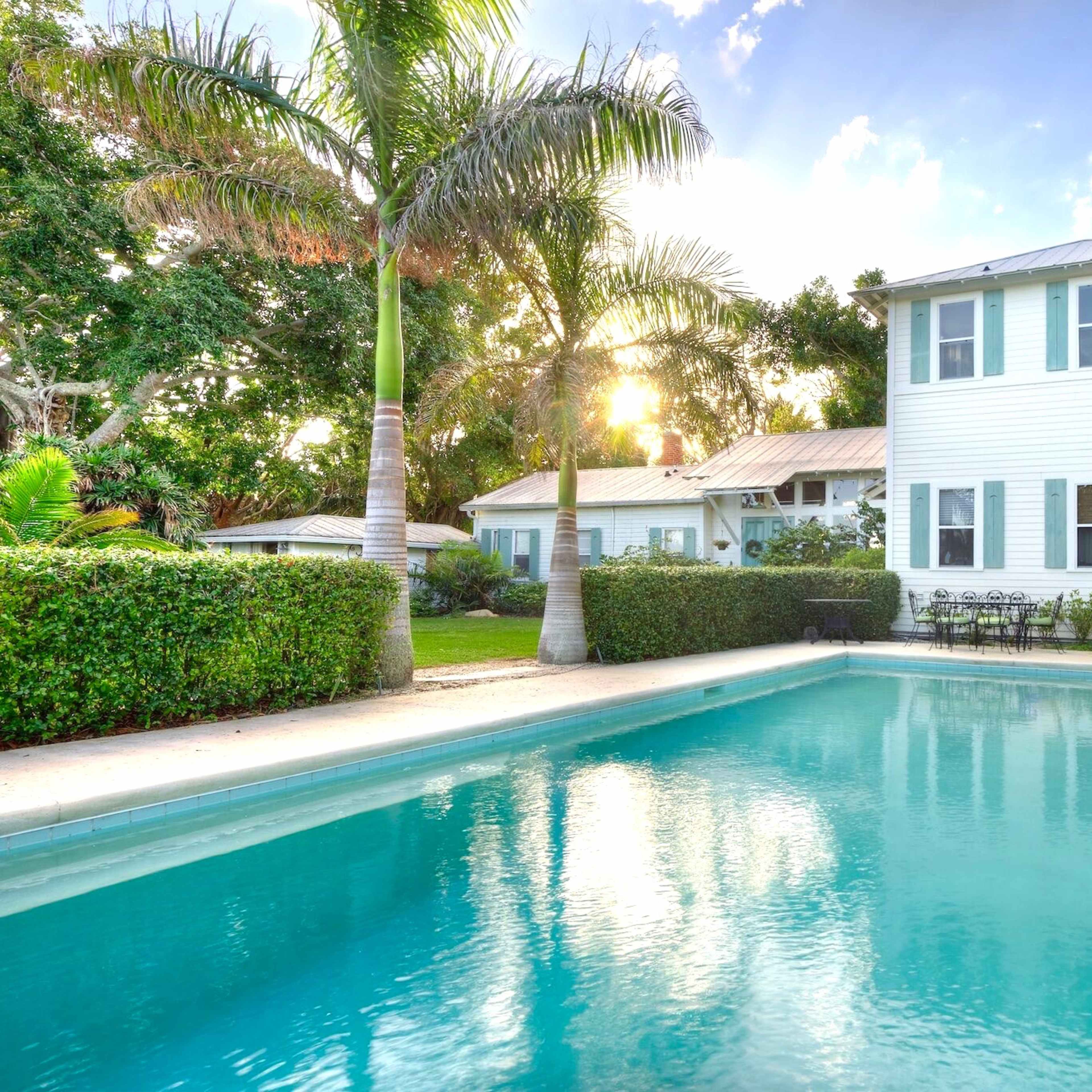 A two-story white house with large windows overlooks a clear swimming pool surrounded by palm trees and landscaped greenery.